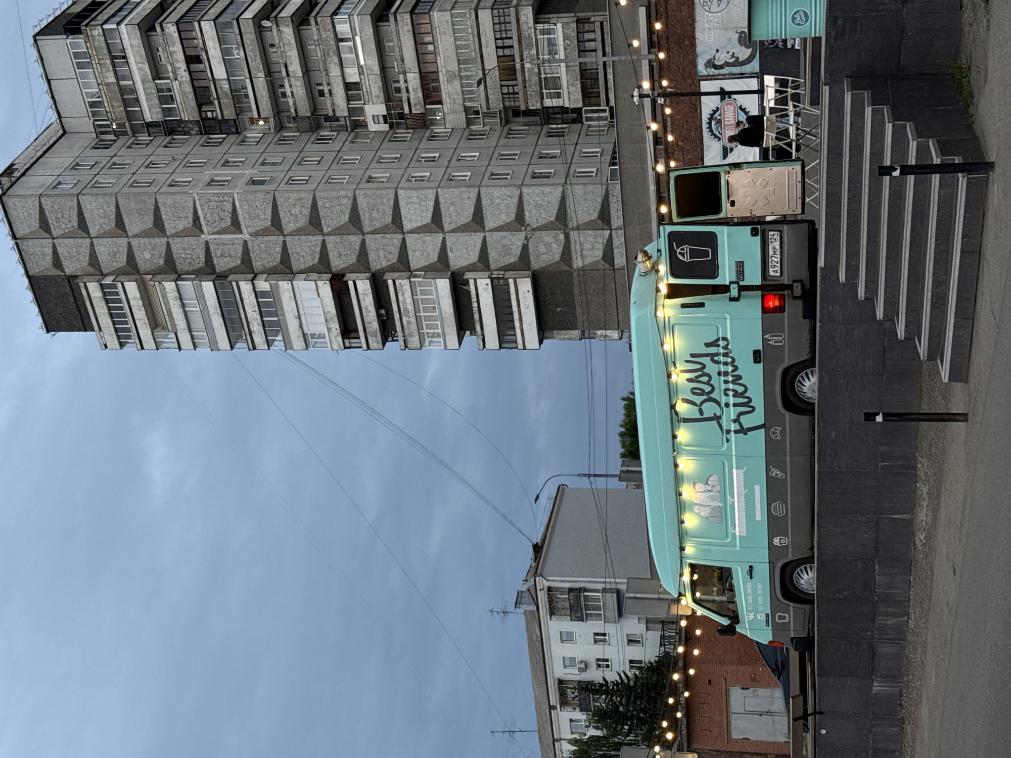The image depicts a food truck with the branding "Best Friends" parked in front of a tall, brutalist-style apartment building. The food truck is turquoise and illuminated by string lights. Several people are visible near the food truck, including two individuals standing and one person sitting at a small table outside. To the right of the food truck, there appears to be a counter or another food service setup. In the background, there is another apartment building and a brick structure. The scene is set outdoors, likely in an urban environment, under an overcast sky. This location is identified as being in the city of Krasnoyarsk, Russia.