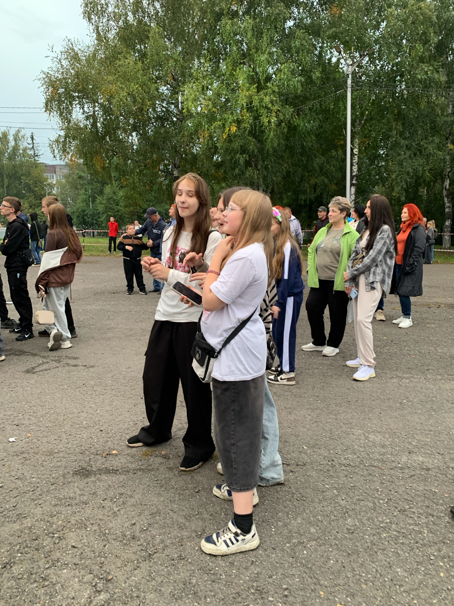 The image shows a group of people standing on an asphalt surface, possibly a parking lot or a schoolyard, in Prokopyevsk, Russia. The background consists of trees with green and yellow foliage, suggesting it is autumn. A building and a street lamp are also visible.
In the foreground, several individuals are captured in various poses. On the left, a person in a black hooded sweatshirt and black pants stands with their arms crossed. To their right, a young person wearing a brown jacket over a white shirt and light-colored pants is holding a small, light-colored purse. Further to the right, two teenage girls are the central focus. One is wearing a white long-sleeved t-shirt with a graphic print and black cargo pants, holding a smartphone in her hand. The other girl has light brown hair and is wearing glasses, with a beaded bracelet on her wrist. She appears to be looking upwards and slightly to the right.
Other individuals in the mid-ground include a person in a green jacket, a woman with dark hair and a plaid shirt, and a woman with red hair wearing an orange top and a black coat. The people are casually dressed, suggesting an informal gathering or event. The lighting indicates an overcast day.