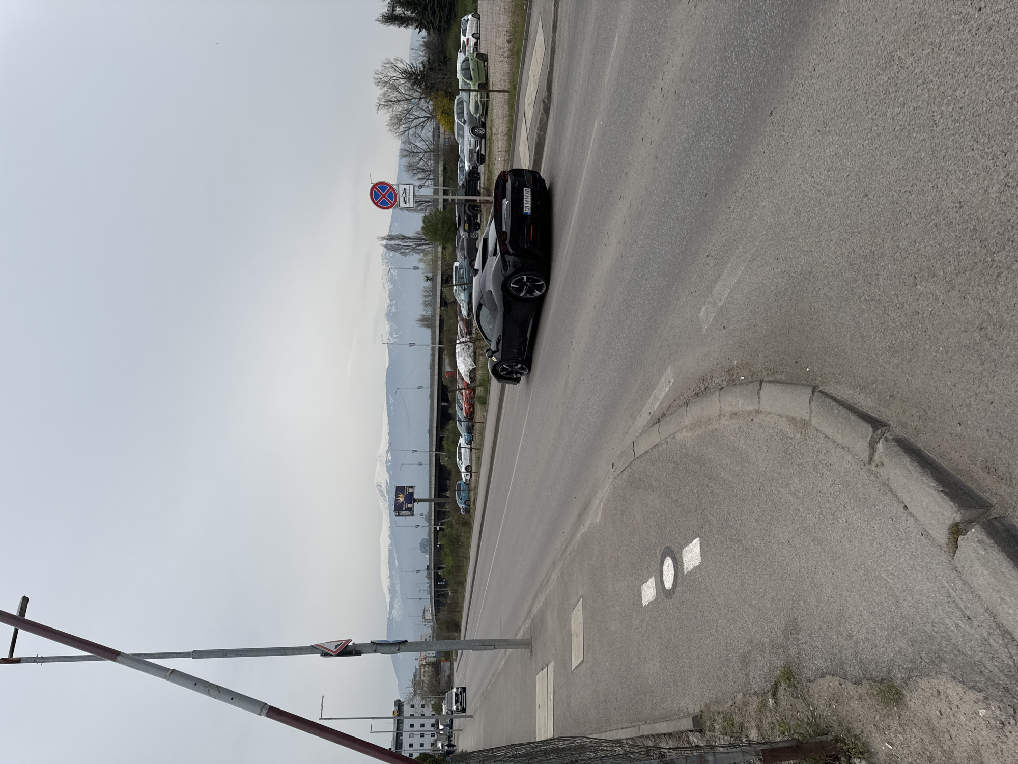 The image depicts a street scene in Sofia, Bulgaria. A black car is parked on the side of the road next to a no-stopping sign. In the background, there is a mountain range and a parking lot with several parked cars. There is a railway crossing with a barrier in the foreground. The sky is overcast. The road is asphalt and includes road markings. Buildings can be seen in the distance.
