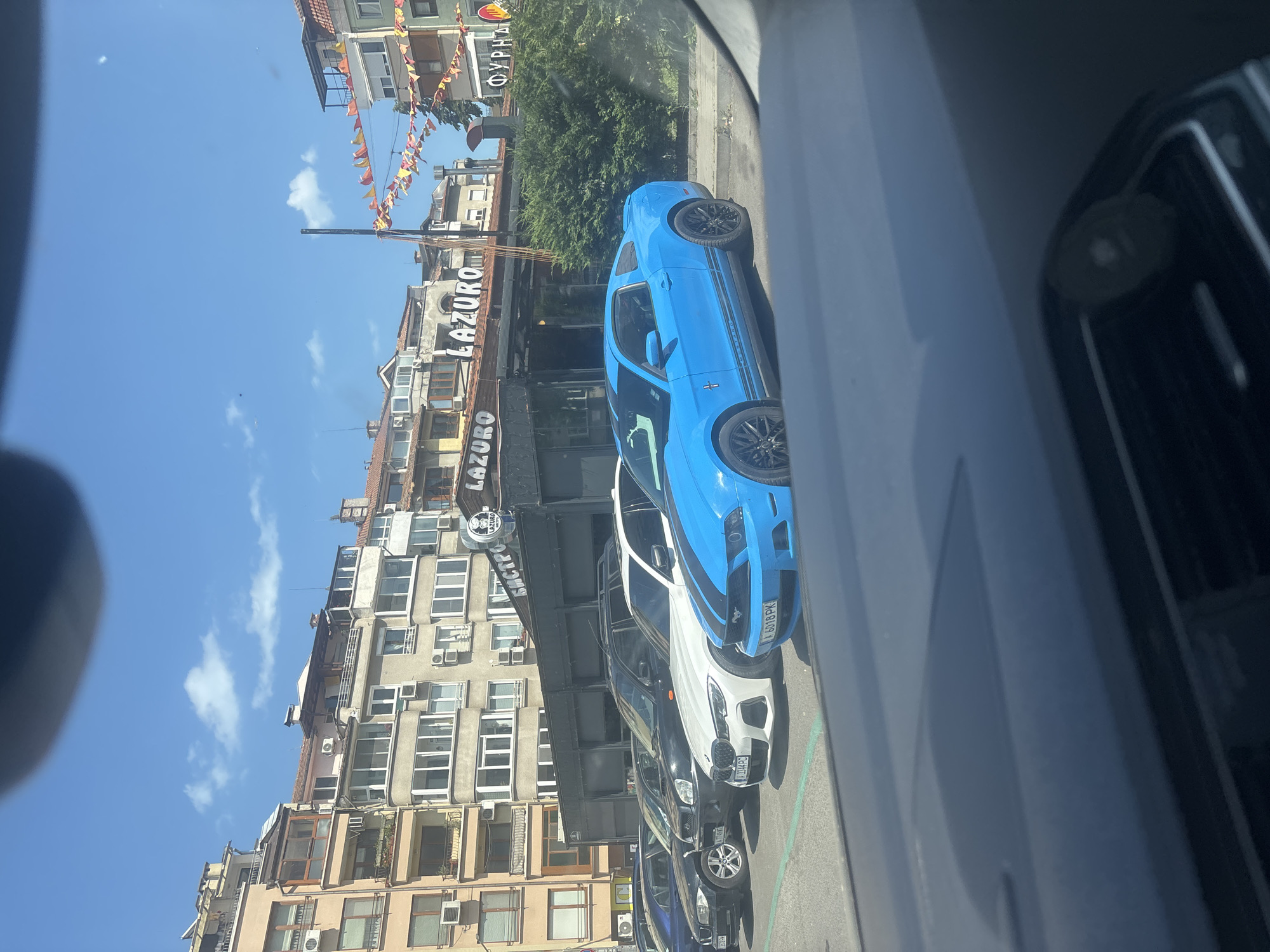 A bright blue Ford Mustang is parked in a lot on a sunny day, with a row of other cars in front of it. Behind the cars, buildings with multiple floors and balconies are visible, along with a restaurant or cafe with the name "LAZURO" displayed. Colorful festive flags are strung across some of the buildings. The scene appears to be in an urban setting.