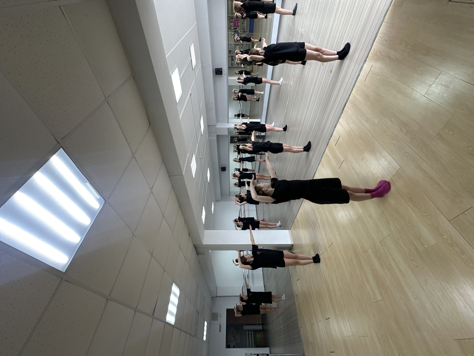 This media file captures an indoor training or dance studio where a group of young people, primarily female, are engaged in a structured exercise session. The setting features a prominent mirrored wall on the right, reflecting the participants and the ceiling, giving the impression of a larger space.
The main subjects are numerous individuals, mostly dressed in black athletic tops and black shorts. They are performing a similar pose, lying on their backs with arms extended overhead and legs stretched out, indicative of a core strengthening or conditioning exercise, possibly a "hollow body" hold or part of a warm-up routine for dance, gymnastics, or fitness. Some individuals are actively holding the pose, while others appear to be resting or observing. One person in the foreground stands out with bright pink socks, while others wear black or white.
The room has a light-colored wooden floor with visible seams between planks. The ceiling is made of white acoustic tiles arranged in a grid pattern, interspersed with bright, rectangular fluorescent light fixtures that illuminate the space. The artificial lighting suggests an indoor activity, and the overall environment is functional and well-lit.
A notable detail is a white rectangular sign on the back wall with visible text: "ВЫХОД" (VYKHOD), which is Russian for "EXIT," consistent with the provided location context of Votkinsk, Russia. The participants appear focused on their activity, demonstrating concentration and discipline.