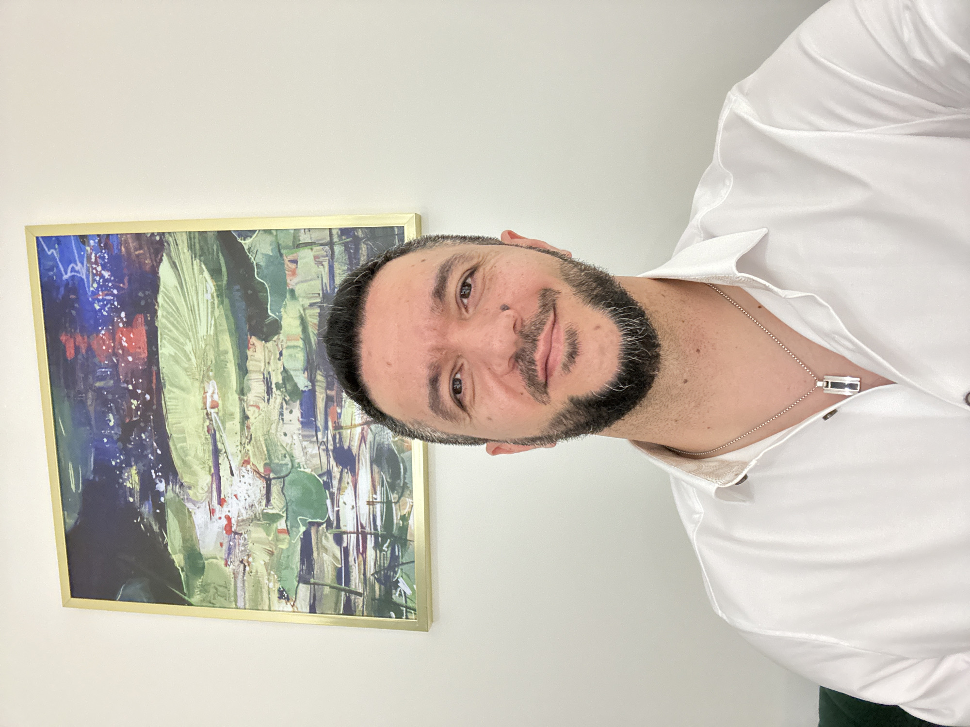 A man with short dark hair and a trimmed beard is pictured indoors, wearing a white button-down shirt and a silver chain necklace with a rectangular pendant. He is facing the camera with a slight smile. To his left is a framed abstract painting with a gold frame and a mix of colors including blues, greens, reds, and whites. The wall behind him is a neutral color, possibly light beige. The location within Warsaw, Poland, cannot be determined from the image.
