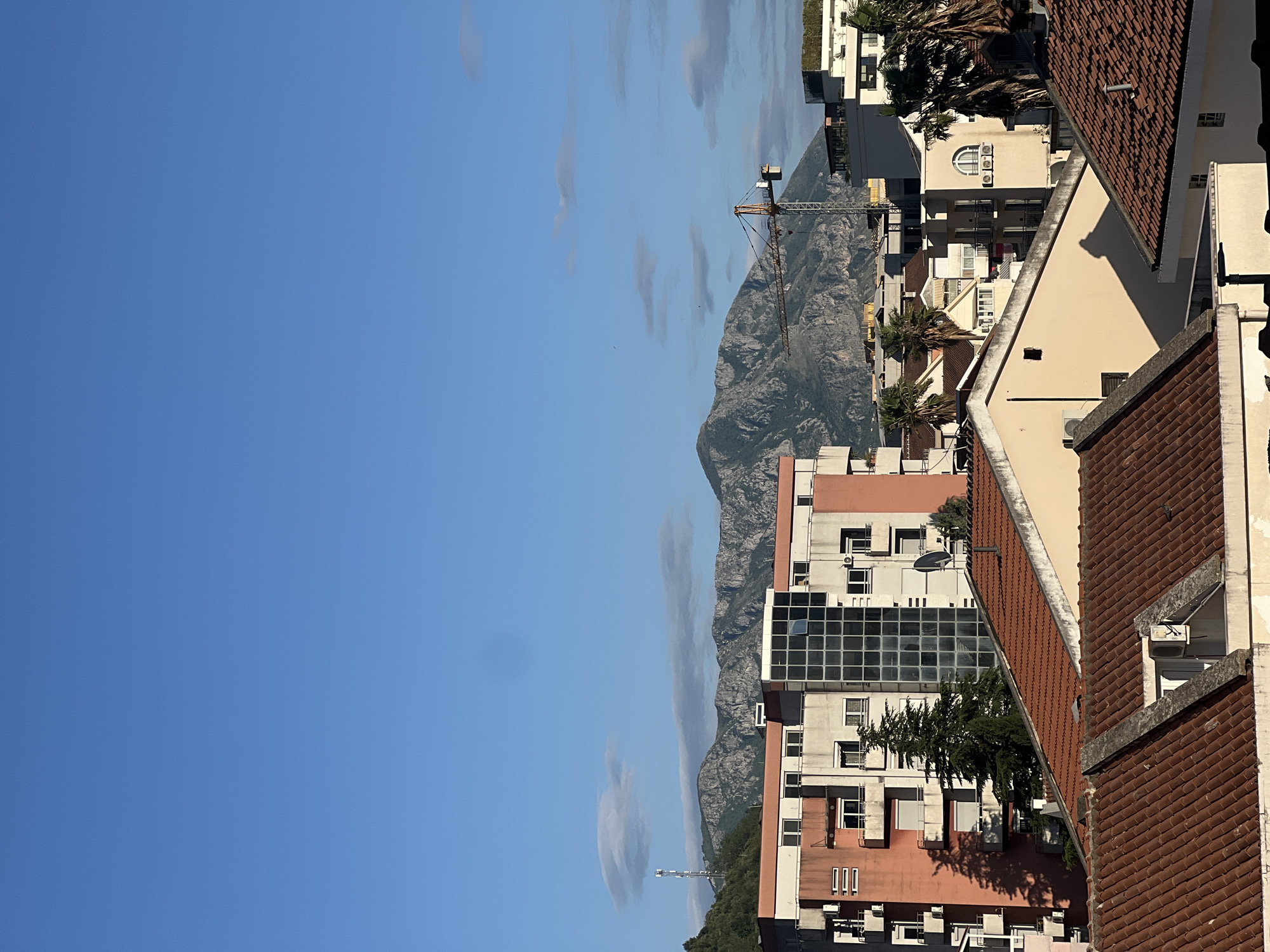This image presents a panoramic view of an urban area in Budva, Montenegro, set against a majestic mountain backdrop under a bright blue sky.
In the foreground and midground, a dense collection of multi-story buildings dominates the scene. These structures exhibit a variety of architectural styles, including modern buildings with large, dark-framed windows, and more traditional residential blocks featuring beige, peach, and white facades with numerous balconies and external air conditioning units. Many roofs are covered with characteristic reddish-brown terracotta tiles, a common sight in the Mediterranean region. Interspersed among the buildings are several tall palm trees, contributing to the coastal atmosphere. The sunlight casts clear shadows, suggesting it is daytime, likely mid-morning to early afternoon.
Above the buildings, a rugged, grey mountain range stretches across the horizon, forming a dramatic natural boundary to the cityscape. Its rocky peaks and undulating contours are clearly visible.
A prominent yellow and black construction crane stands tall against the mountains, indicating ongoing development within the city. On the far left, a slender communication tower can also be seen on a lower hill. The sky overhead is a vibrant clear blue with a few sparse, elongated white clouds, suggesting fair weather. There is no discernible human activity or visible text in the image.