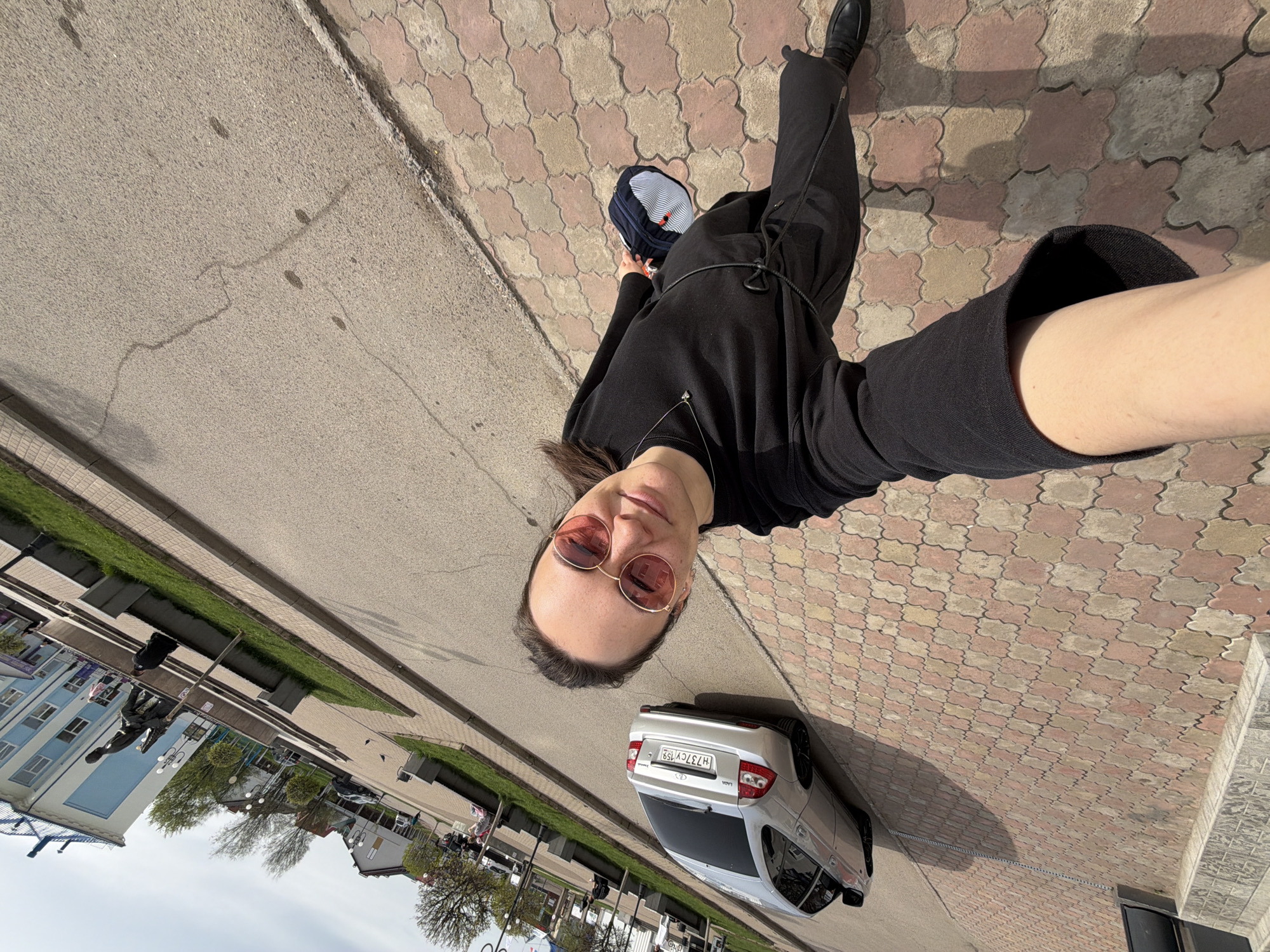 A person, likely female, is taking a selfie, rotated counter-clockwise, on a paved street in Chaikovsky, Russia. The person has brown hair, is wearing round sunglasses with rose-colored lenses, a black hooded sweatshirt, black shorts or a short black dress, black leggings, and black shoes. They are holding a blue and white striped bag in their left hand. A silver car is parked behind them, with the license plate H37CY 159 clearly visible. The street consists of gray concrete and patterned pavement. In the background are buildings, trees, and street furniture. The sky is overcast.