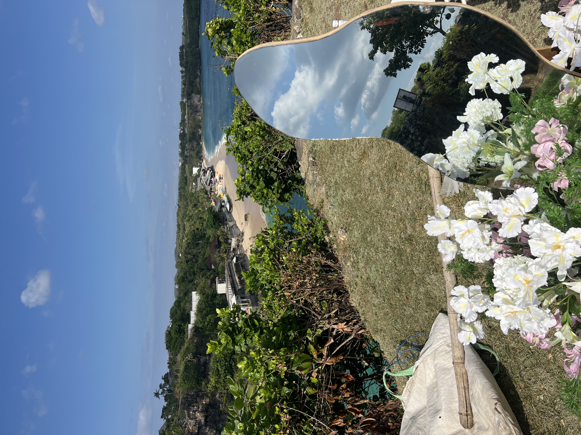 The image captures a vibrant, sunlit panoramic view of a tropical coastline in Kuta Selatan, Indonesia, seen from an elevated perspective, likely a cliff. In the foreground and midground, a large, irregularly shaped mirror with a light-colored frame is prominently featured. The mirror reflects a brilliant blue sky dotted with fluffy white clouds, along with surrounding green foliage and a faint, dark sign containing unreadable text. Adorning the right side and bottom right of the mirror are arrangements of white and light pink flowers, suggesting a decorative setup or photo opportunity. A bamboo pole rests on a light-colored canvas or burlap bag amidst the flowers in the lower right.
Beyond the mirror and foreground grass, the stunning backdrop unfolds. A sandy beach curves along the coastline, meeting the clear, turquoise-blue ocean. Small waves gently break on the shore, and several boats or beach structures are visible on the sand. The cliffs and surrounding landscape are densely covered with lush, vibrant green tropical vegetation. The bright sunlight and clear sky indicate a midday or early afternoon setting with excellent weather conditions. No people are visible in the scene.