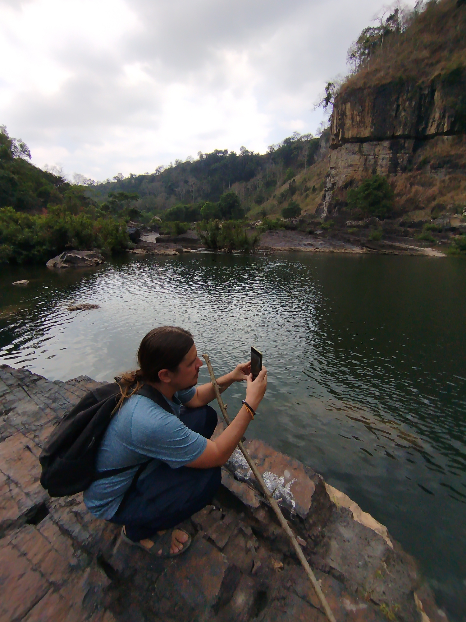 A person with long brown hair is squatting on a rocky ledge next to a body of water, holding a smartphone and taking a photograph. They are wearing a blue t-shirt, dark pants, and sandals, and have a black backpack. A wooden stick rests on the rocks beside them. The body of water is surrounded by lush greenery and a rocky cliff face. The sky is overcast with grey clouds. The location is inferred to be near Tân Phú, Vietnam, based on general landscape characteristics.