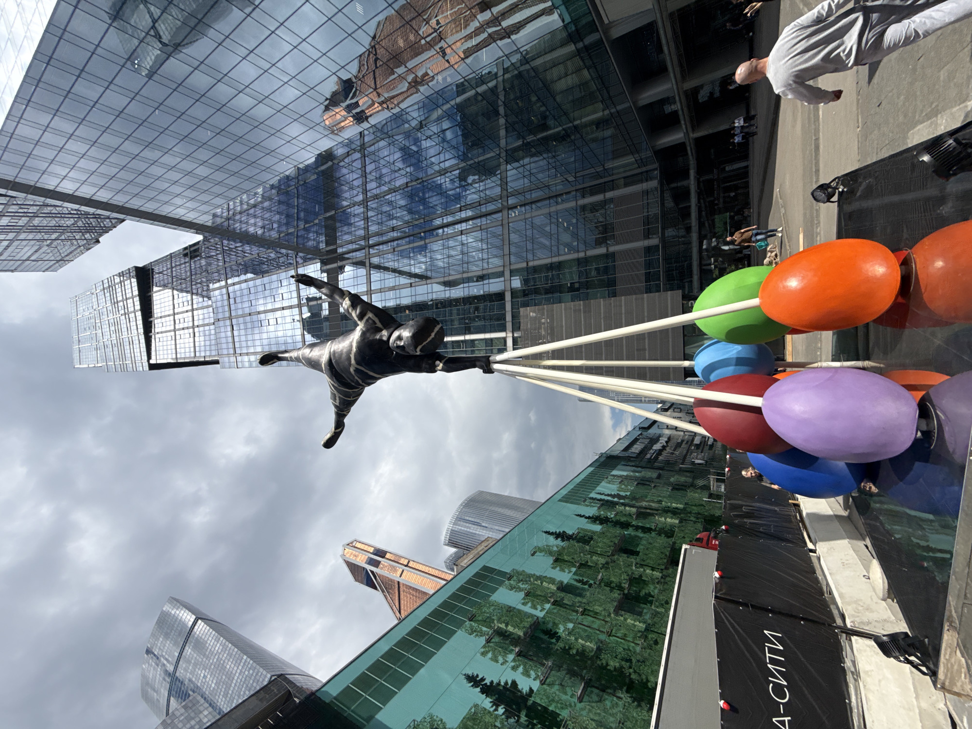 In Moscow, Russia, a low-angle, wide shot captures an outdoor public space dominated by modern skyscrapers. In the foreground, a sculpture of a human figure in a handstand, made of dark material, is supported by three white poles. Below the sculpture, several large, colorful, egg-shaped objects are arranged: a red one, a purple one, two blue ones, a green one, and an orange one. Reflections of the surrounding architecture and sky are visible on the polished surface beneath the eggs.
To the left, a building with a green facade featuring a printed image of a forest is visible. A large grey truck is parked near a black barrier with white text reading "A-СИТИ". In the background, a cluster of sleek, glass-faced skyscrapers forms the urban backdrop. Some people are seen walking in the distance. A man in a grey tracksuit is walking away from the camera on the right side of the frame, and another man in a brown shirt is walking towards the left in the middle ground. The sky is overcast with scattered clouds.