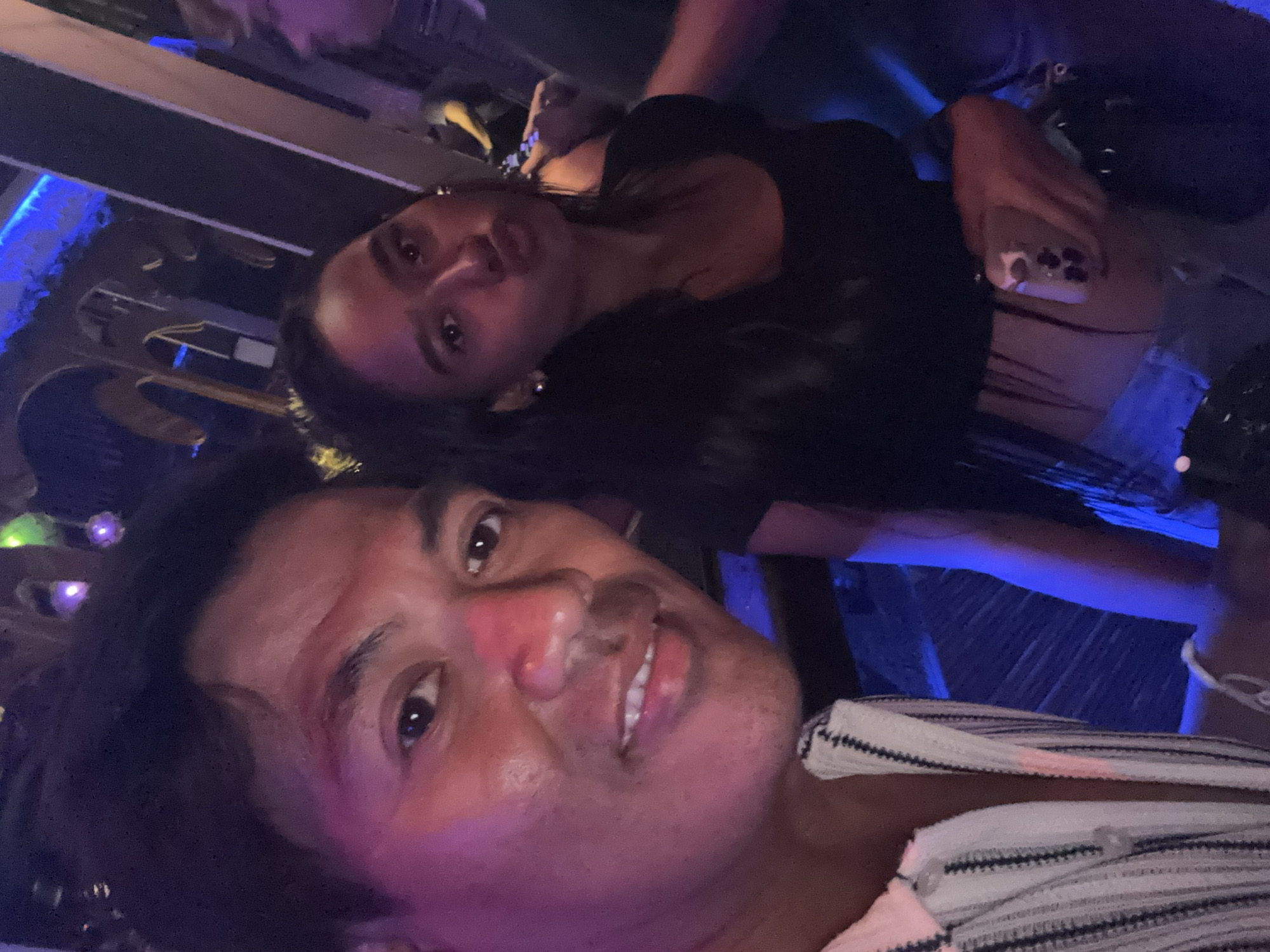 The image shows a close-up selfie of two individuals, a man and a woman, in what appears to be a dimly lit, indoor establishment. The man, on the left, is smiling and looking directly at the camera. He is wearing a striped shirt. The woman, on the right, is pouting and holding a smartphone, suggesting she is taking the photo. She is wearing a black top and denim shorts. The background is blurry, but it seems to be a bar or club with blue lighting and some decorative elements, possibly in North Kuta, Indonesia, given the context.