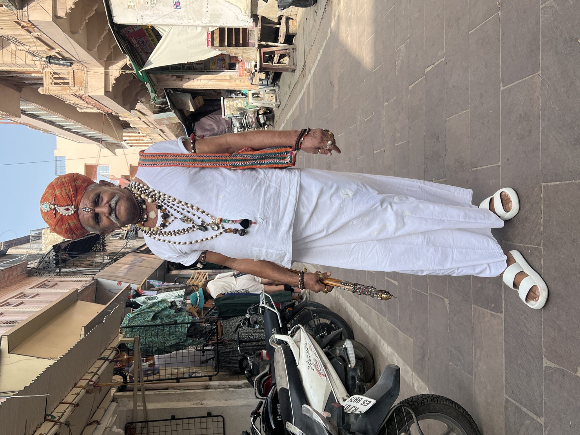 An elderly Indian man, likely a local resident of Pushkar, India, stands on a paved street. He wears a traditional white kurta and dhoti, adorned with multiple beaded necklaces and bracelets. A vibrantly orange turban, embellished with intricate beadwork, crowns his head, and a prominent mustache completes his distinguished appearance. In his right hand, he holds a decorative staff. The background reveals a bustling street scene with shops and motorcycles, indicative of a vibrant Indian marketplace. The architectural style of the buildings suggests a historic urban setting, possibly within the religious city of Pushkar.