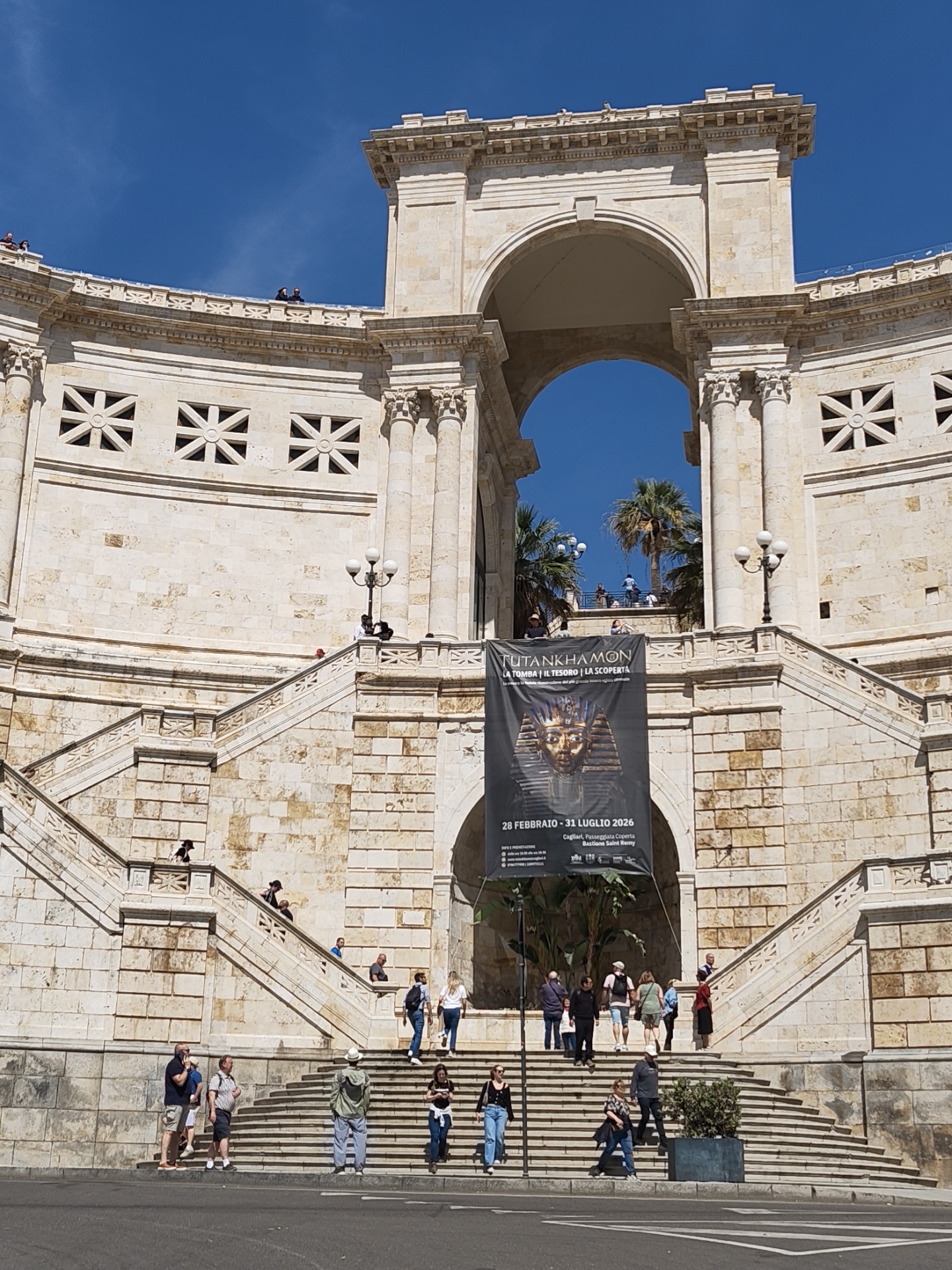 The image shows the Bastione di Saint Remy in Cagliari, Italy. The bastion is a large, white stone structure with arches and stairs leading up to the top. A large banner advertising a "Tutankhamon" exhibit is hung over the central archway. People can be seen walking up and down the stairs. The sky is blue. The banner reads "Tutankhamon la Tomba il Tesoro la Scoperta" and "28 Febbraio-31 Luglio 2026" and "Cagliari, Passeggiata Coperta Bastione Saint Remy".