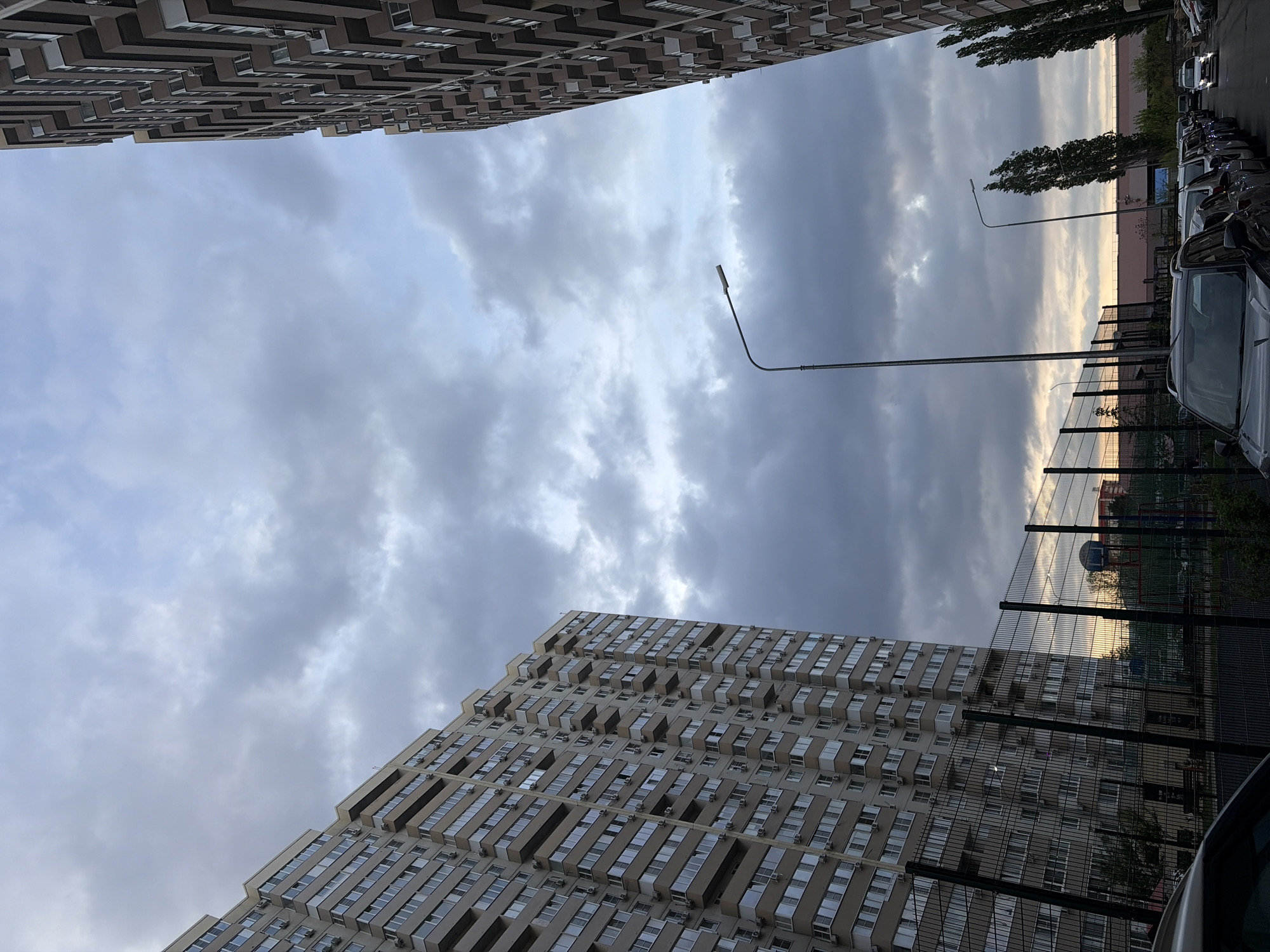 The image is a low-angle shot of a street scene in Tliustenkhabl', Russia. Two tall apartment buildings frame a view of a cloudy sky. A row of parked cars lines the street, separated from a fenced-in area, possibly a park or playground. Streetlights are visible extending into the sky. The overall impression is a residential area on a cloudy day.