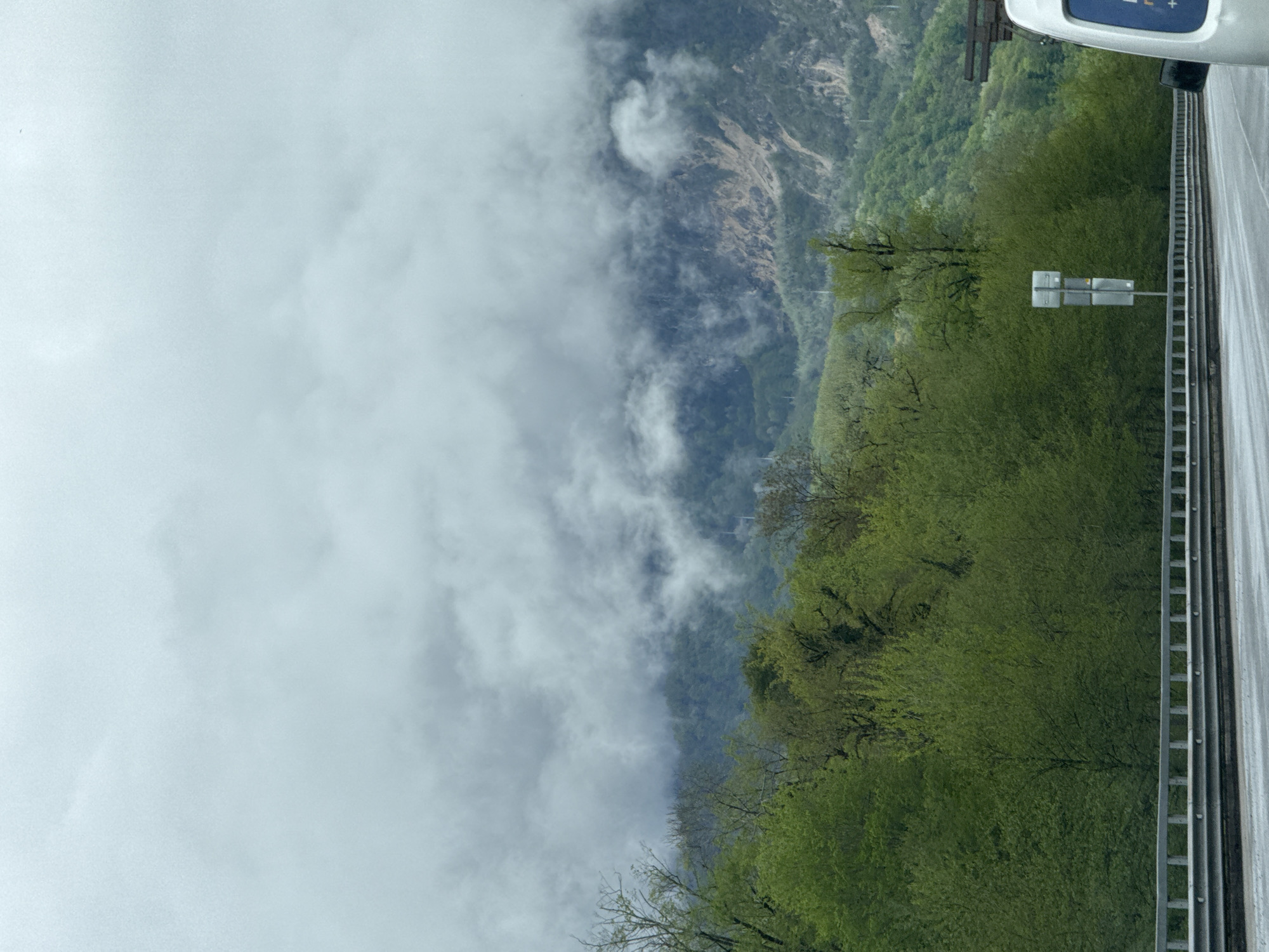 The image captures a roadside view in what appears to be a mountainous region in the city of Adlerskiy, Russia. On the right side of the frame is a highway road with a metal barrier along the edge. Dense green trees and shrubbery line the roadside and obscure views. Beyond the foliage, a mountain range is partially shrouded in clouds. The sky is filled with thick, gray clouds, suggesting overcast weather conditions. A portion of a vehicle's exterior is visible at the top right corner of the image.