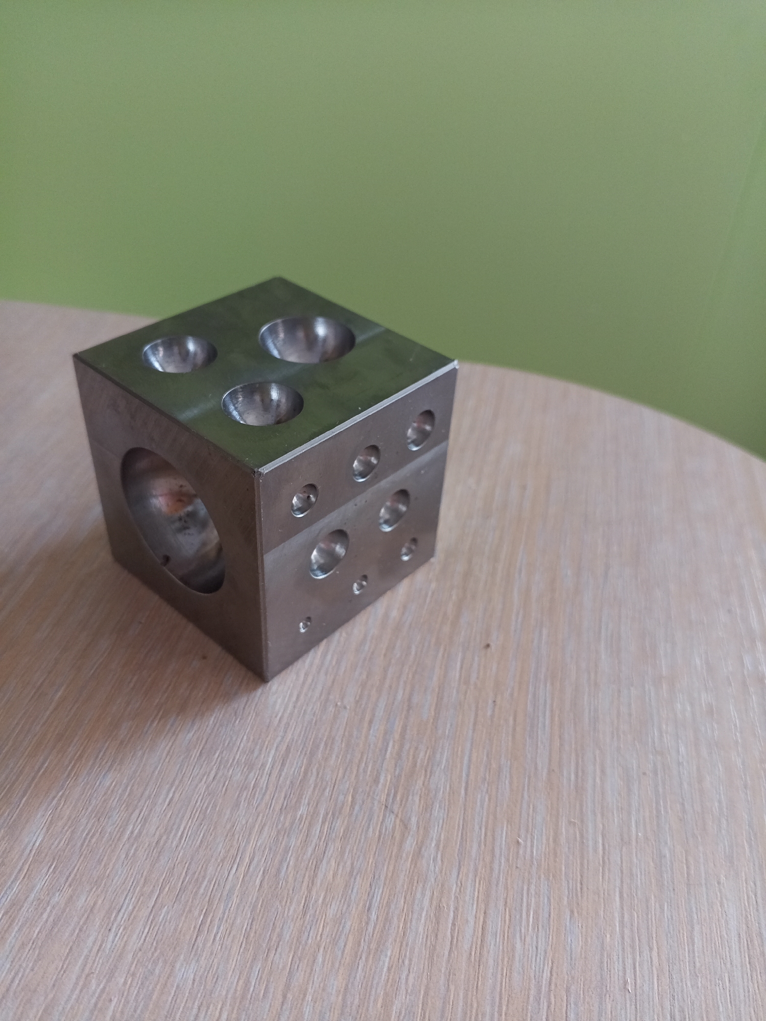 A metal cube with various indentations and holes sits on a light-colored wooden surface. The cube is positioned in the lower left quadrant of the image. The background is a solid, light green color. The image is taken from a slightly elevated angle looking down. The scene appears to be indoors, likely on a table. There is no indication of the city of Vilyuysk, Russia in the image.