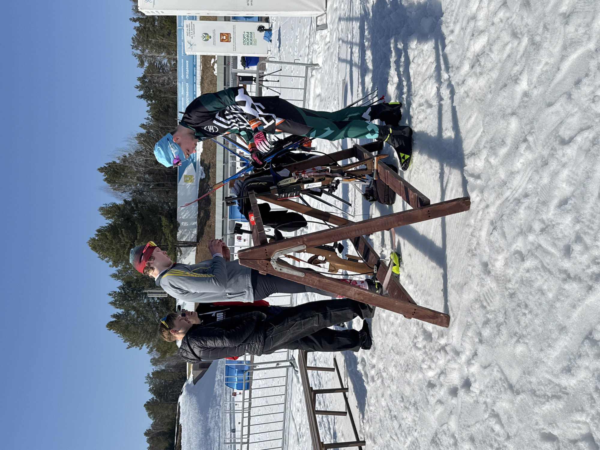 This image depicts an outdoor winter sports scene in Прикамский, Russia, under a clear blue sky on a sunny day, with long shadows cast across the snow-covered ground. Four males are present in the foreground.
On the far right, a male wearing a black, green, and white biathlon suit, a light blue cap, pink-rimmed sunglasses on his head, and black ski boots with yellow accents, is actively engaged with equipment on a central wooden A-frame rack. This rack holds multiple pairs of skis, identifiable brands include Atomic and Fischer, along with several ski poles and at least two biathlon rifles, one featuring a prominent wooden stock.
To his immediate left, a male wearing a grey long-sleeved shirt with yellow stripes, a red headband, and yellow-framed sunglasses on his head, is looking down at an item held in his hands. Further to the left, two additional males stand. One is dressed in a black puffer jacket with sunglasses on his head. The other is wearing a black and white PUMA hoodie, also with sunglasses on his head; his face is partially obscured.
In the background, a metal barrier fence separates the foreground from the mid-ground. A wooden bench, partially covered by snow, is visible to the left of the fence. Behind the fence, white banners display Cyrillic text, including "СПОРТ НОРМА ЖИЗНИ" (Sport is the Norm of Life) and the Russian coat of arms, alongside a small blue structure. A dense line of pine trees forms the horizon, with large snow banks visible in the far background. The snow on the ground shows visible footprints and a textured surface.