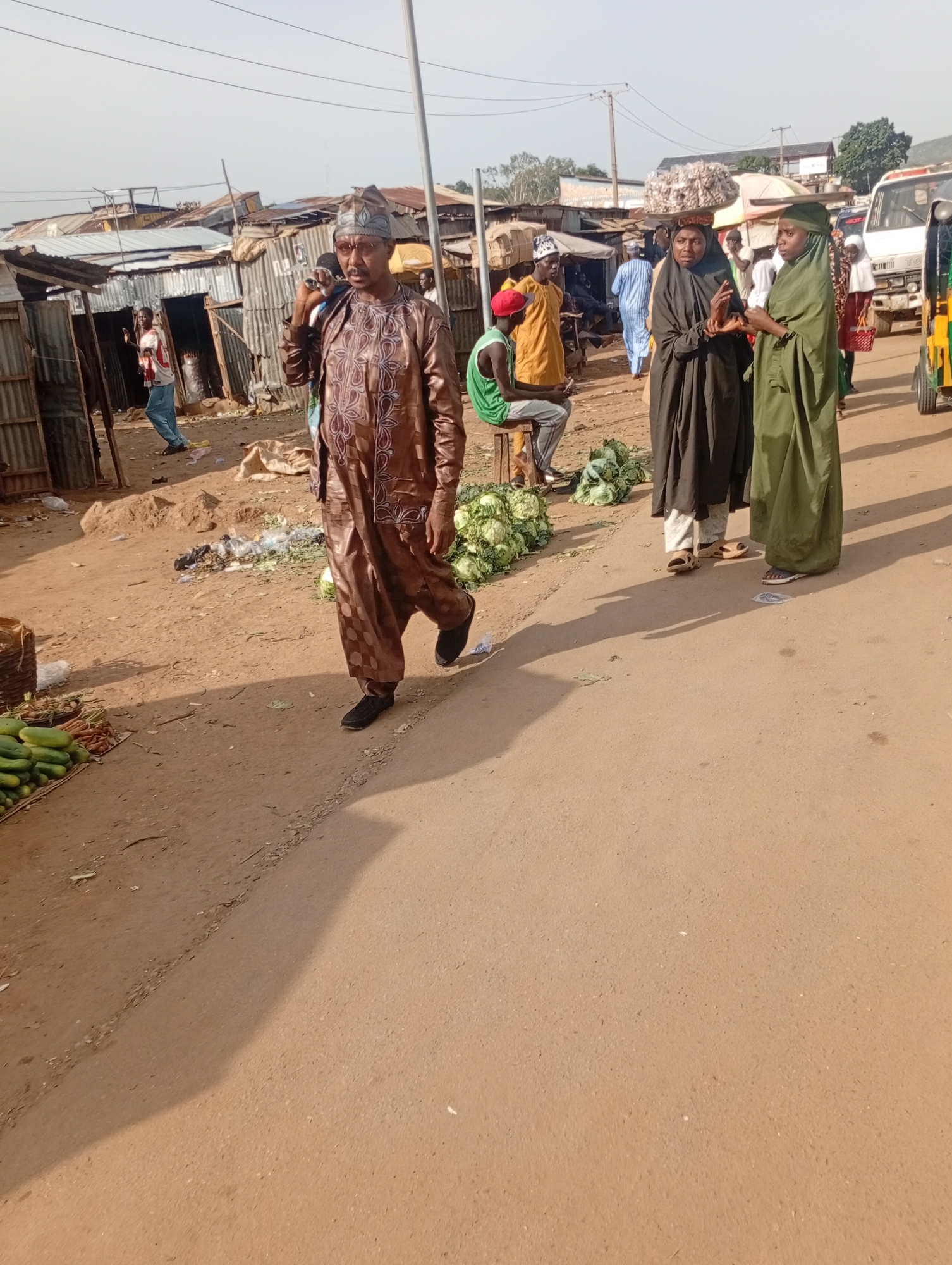 In Rafin Par, Nigeria, an outdoor marketplace is captured on a clear day. The foreground features a man in a brown embroidered robe and cap, walking along a dirt path that transitions into a paved road. To his left, a display of fresh vegetables, including cucumbers and carrots, is arranged. In the background, several structures made of corrugated metal and wood line the street, suggesting informal market stalls. People are visible in the midground, including women wearing hijabs, one carrying a basket of goods on her head, and men in casual attire. A white truck and a colorful three-wheeled vehicle are parked further down the road, indicating ongoing commercial activity. The overall scene depicts a bustling, everyday market environment.