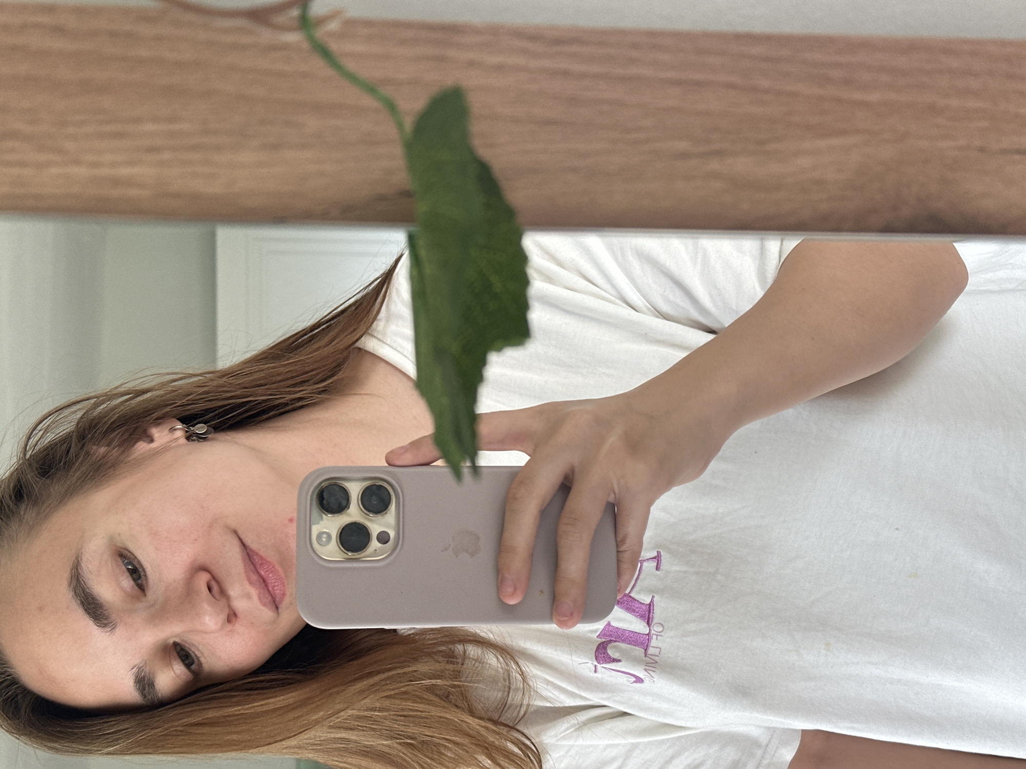 A woman is taking a selfie in a mirror, holding a smartphone in her right hand. She has fair skin, light brown hair, and is wearing a white t-shirt. A green leaf is partially obscuring her face. The background appears to be a room with a wooden shelf or ledge visible above the mirror. There is no information suggesting this photo was taken in Kharnavy, Russia.