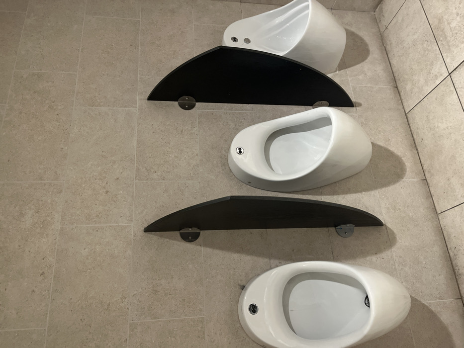 The image presents a modern public restroom interior, specifically a wall-mounted urinal arrangement, located in Dorno, Italy. Three sleek, white, teardrop-shaped urinals are mounted vertically on a light-colored tiled wall. Each urinal features a small, metallic flush mechanism or sensor with two circular elements.
Between the urinals are two dark, semi-circular shelves, also mounted on the wall. These shelves appear to be made of a dark, possibly wooden, material and are supported by two visible metallic brackets each. The wall itself is covered in rectangular, light beige tiles with a subtle, textured finish, laid in a horizontal pattern. Similar tiles are visible on the floor, suggesting a uniform aesthetic.
The scene is illuminated by an unseen light source, casting gentle shadows from the urinals and shelves onto the wall, emphasizing their form and depth. No people are present, and no activity is occurring; the urinals are empty and appear clean and well-maintained. The contemporary design of the fixtures and the clean tiling indicate a modern and functional public facility. There is no visible text in the image.