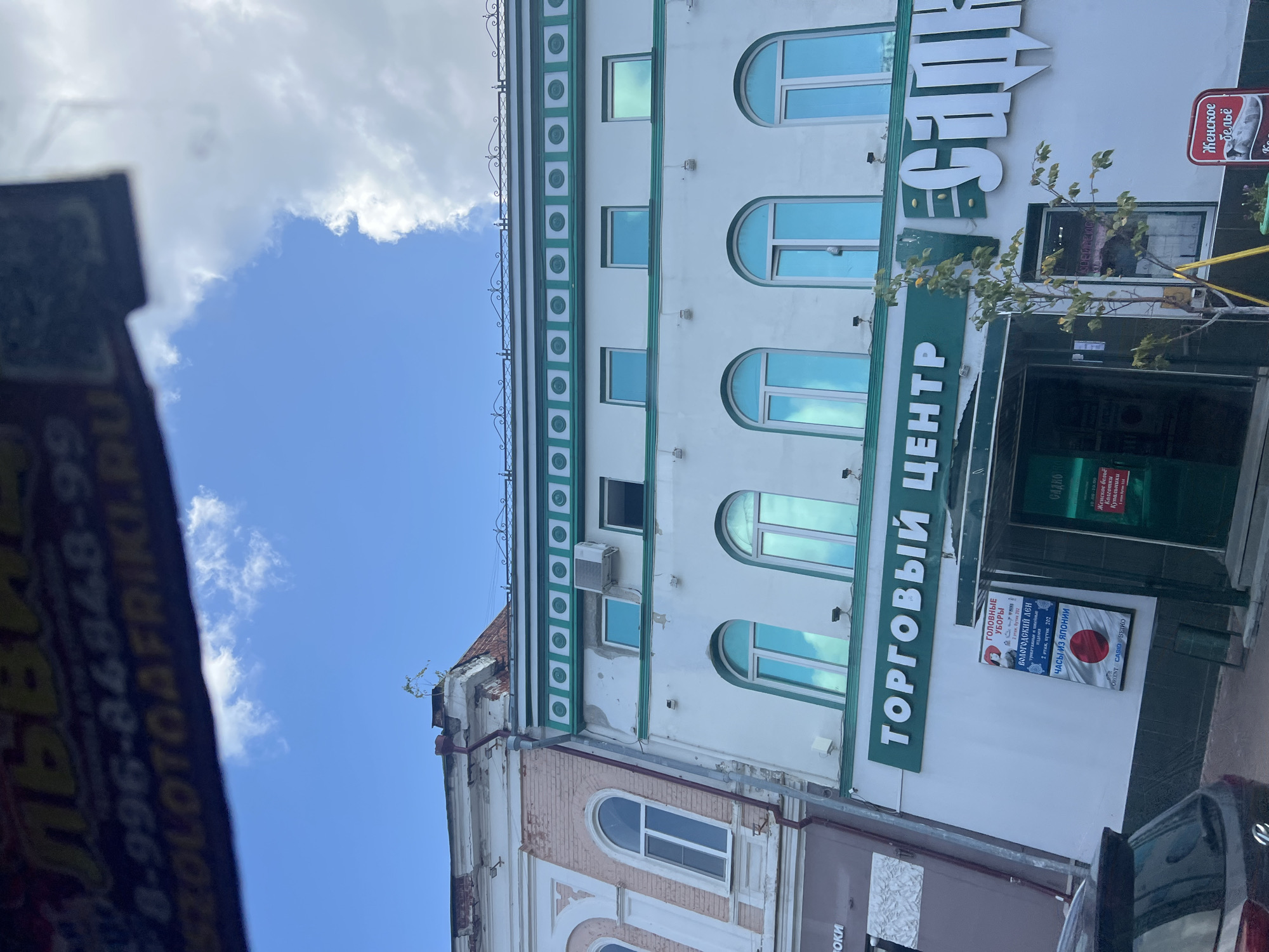 The image captures a street view in Ulyanovsk, Russia, focusing on the facade of a commercial building. The building has a white exterior with teal green decorative accents and trim around the windows and roofline. Several arched windows with teal blue reflective glass are visible on the upper floors. A prominent green sign above the entrance reads "ТОРГОВЫЙ ЦЕНТР" (Trading Center) in white lettering. To the right of this, a large, stylized white sign partially visible reads "САД" (Garden). Below the "ТОРГОВЫЙ ЦЕНТР" sign, there are two smaller signs. The left sign, predominantly blue, advertises "ГОЛОВНЫЕ УБОРЫ" (Headwear) and "ЧАСЫ ИЗ ЯПОНИИ" (Watches from Japan), with details about an address and floor. The right sign, red and white, advertises "Женское бельё" (Women's Lingerie). A small, young tree with green leaves stands to the right of the entrance. In the foreground, the rear of a dark-colored car is visible. The sky above is bright blue with scattered white clouds. The architectural style suggests a mix of older and possibly more modern renovations.