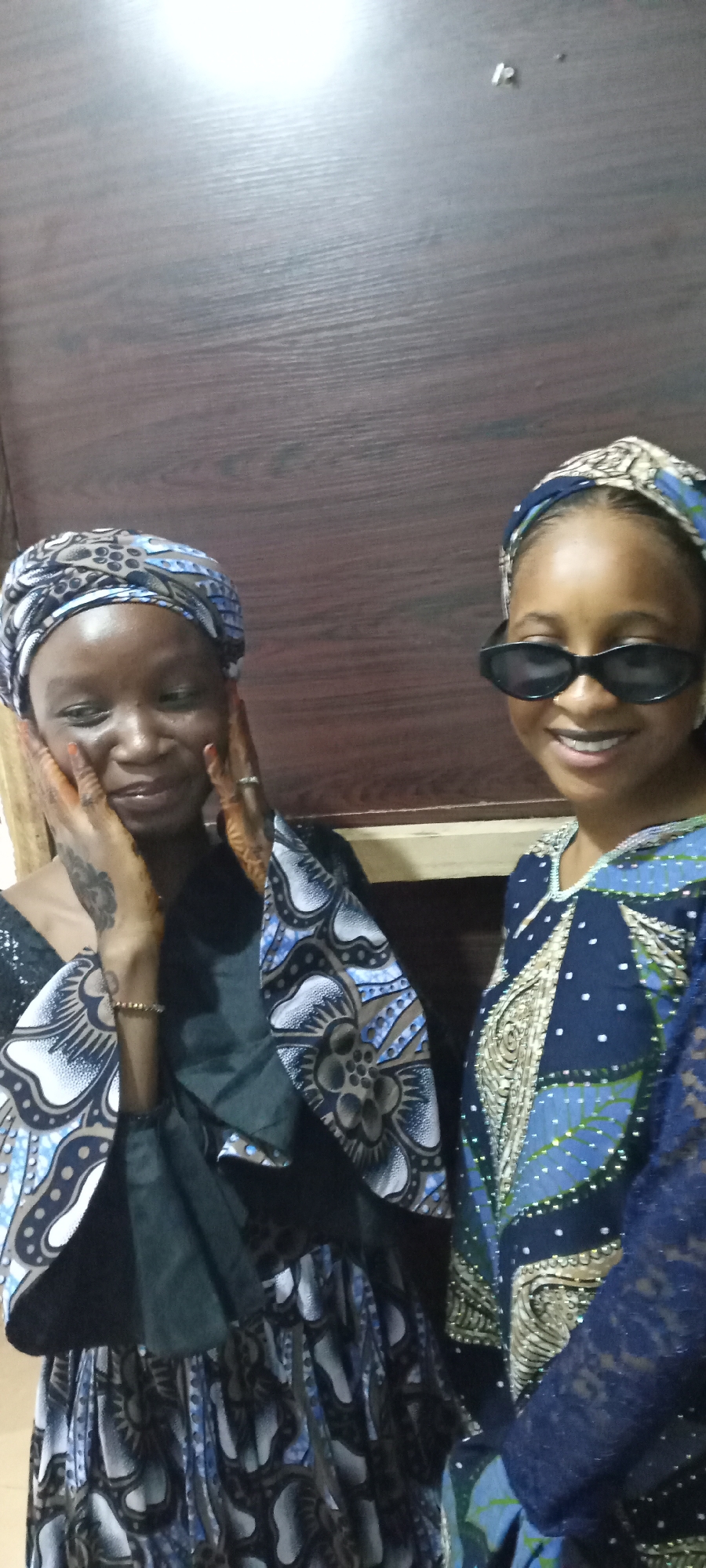Two women are posing for a photo indoors. The woman on the left is wearing a headscarf with a white, brown, and blue pattern. She has a black top with matching patterned sleeves. She is touching her face with her hands, which have henna designs on them. A tattoo is visible on her left hand. On the right, the other woman is wearing a similar headscarf and sunglasses. She has on a blue and white patterned dress with a lace sleeve. The background appears to be a plain wooden wall. The location is in the city of Unknown, Unknown.