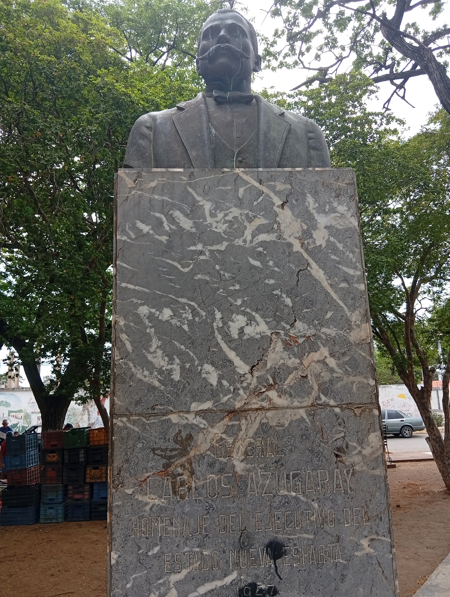 A bronze bust of a man with a prominent mustache, wearing a suit and bow tie, stands atop a marble pedestal. The pedestal is inscribed with text in raised letters. The inscription reads: "GENERAL CARLOS AZUGARAY HOMENAJE DEL EJECUTIVO DEL NUEVA ESPARTA 1947". The bust and pedestal are situated outdoors, with trees and foliage visible in the background. In the lower left portion of the image, stacked plastic crates are piled up, and a few individuals are present, suggesting a market or public gathering area. A car is parked on a street in the background on the right. The environment appears to be a public square or park. The style of the bust and the inscription suggest it is a historical monument. Based on the inscription, this monument is likely located in Nueva Esparta, Venezuela, and commemorates Carlos Azugaray. It is situated in the city of Juan Griego, Venezuela.