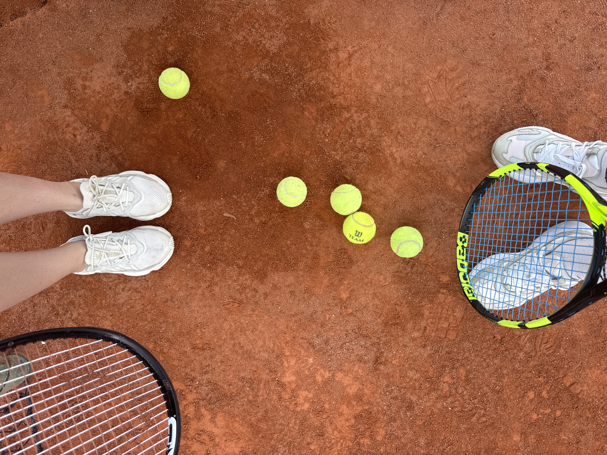 This is an overhead, close-up shot of a clay tennis court in Neubiberg, Germany. The image features two pairs of white tennis shoes and multiple tennis balls scattered across the reddish-brown surface. The court shows faint footprints and scuff marks. Two tennis rackets are visible, one in the foreground with a neon yellow and black frame, and another partially visible at the top left with a black frame. One pair of shoes is positioned near the top of the frame, and the other pair is in the bottom right, partially obscured by the racket. Six tennis balls are visible in total. One ball is near the top right, and the other five are clustered in the center and lower left of the frame. The overall scene suggests an informal game or practice session on a sunny day.