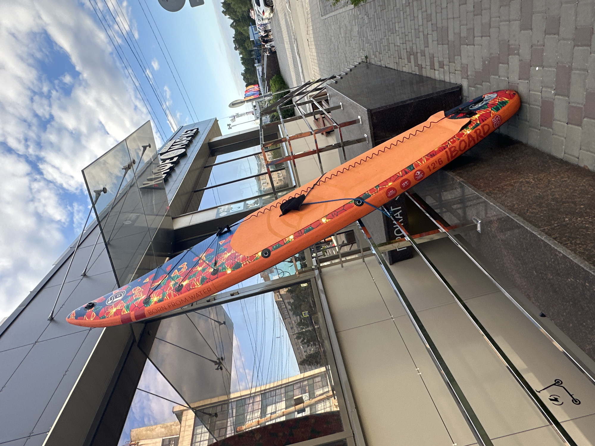 An orange paddleboard with a floral pattern and the text "BORN FROM WATER" and "12'6 IBOARD" is propped against the modern glass-fronted building of a cafe with the name "KAQE MAPKE" visible. The paddleboard is leaning at an angle, occupying a significant portion of the foreground. To the right of the building, a paved sidewalk leads away from the scene, and a metal railing with stairs is visible. In the background, people and parked cars are seen near the cafe entrance, with trees and more buildings in the distance. The sky above is partly cloudy.