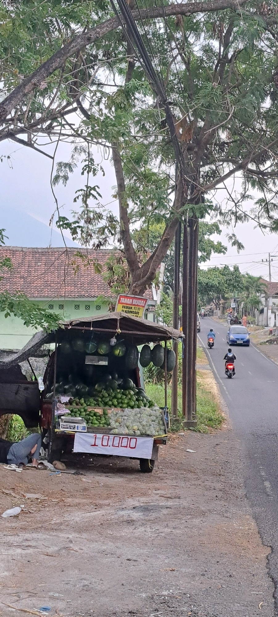A roadside fruit stand is set up on the shoulder of a paved road in Sumber Gedang, Indonesia. The stand, mounted on a three-wheeled vehicle, displays a large assortment of green fruits, possibly avocados, piled high. Several watermelons are also visible, hanging from the roof of the stand and displayed on shelves. A sign with red lettering reads "10.000," indicating the price. To the left of the stand, a person, dressed in dark clothing and blue jeans, is lying down on the ground, appearing to be resting or sleeping. A scale is placed on the stand next to the produce. In the background, a blue car and several motorcycles are traveling on the road. A building with a red tile roof is situated behind the fruit stand. Overhead, a dense network of electrical wires runs through the branches of a large tree. The scene suggests a typical street-side market environment in a rural or semi-urban area of Indonesia.