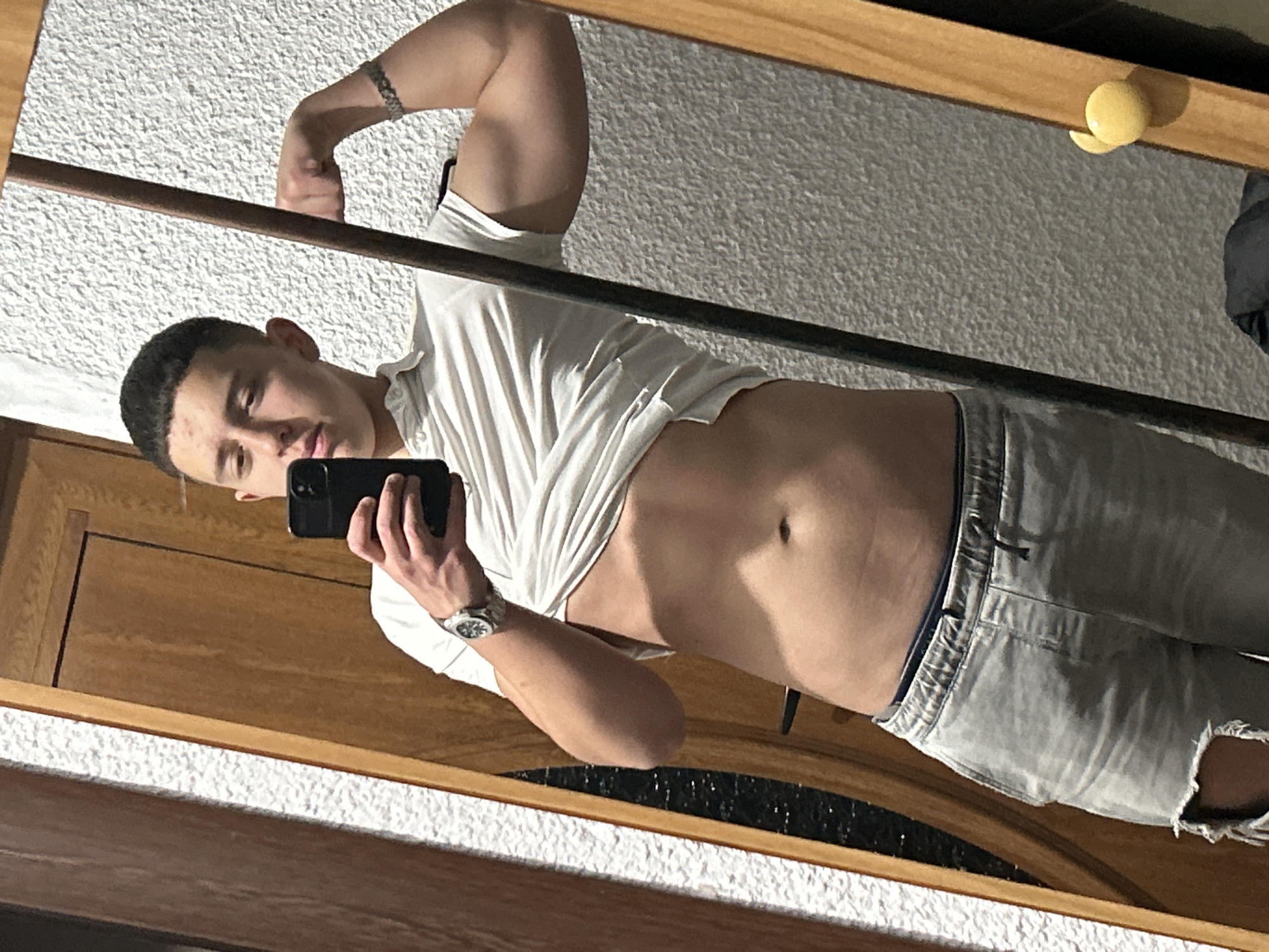 A young man is taking a selfie in a mirror. He is wearing a white shirt that is partially unbuttoned, revealing his abdomen. He is also wearing light-colored jeans with a rip in the knee. He has short, dark hair and a watch on his left wrist. His left arm is flexed, displaying a tattoo on his forearm. The background includes a textured white wall, a wooden frame, and a yellow object in the upper right corner. The location is not evident in the image.