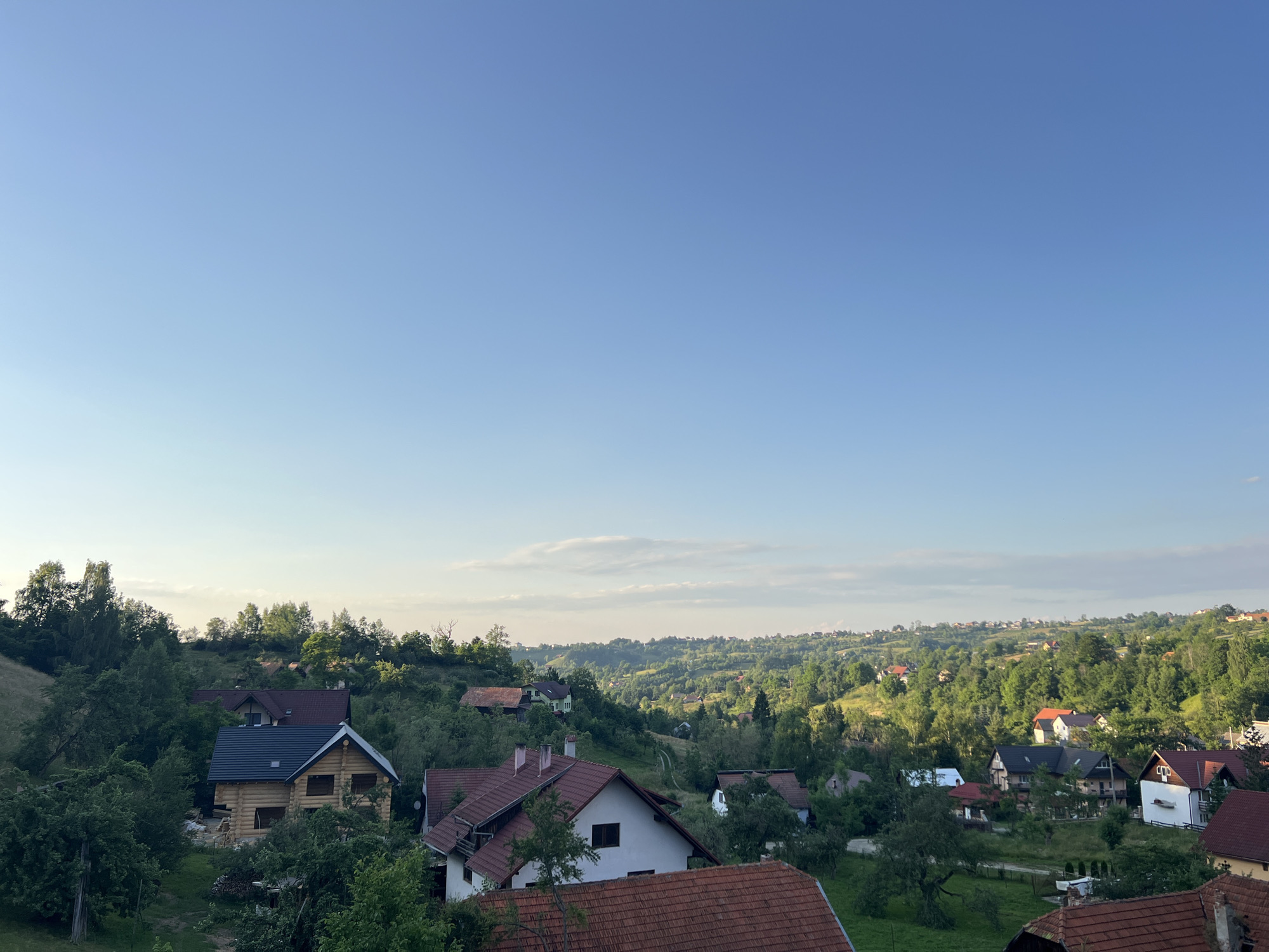 This is a medium-shot, eye-level, horizontal, outdoor photograph of a rural village nestled in rolling green hills under a clear blue sky. The scene depicts a collection of houses, some with red tile roofs and others with darker shingles, interspersed with lush trees and vegetation. A prominent log cabin under construction, with a dark-colored roof, is visible in the mid-ground. The rolling hills extend into the distance, dotted with more houses and trees, creating a sense of depth and a sprawling landscape. The time of day appears to be late afternoon or early evening, given the soft lighting and the clear sky with subtle cloud formations near the horizon. The weather seems pleasant and calm. The overall mood is peaceful and serene, evoking a sense of rural tranquility. No people or text are visible in the image.