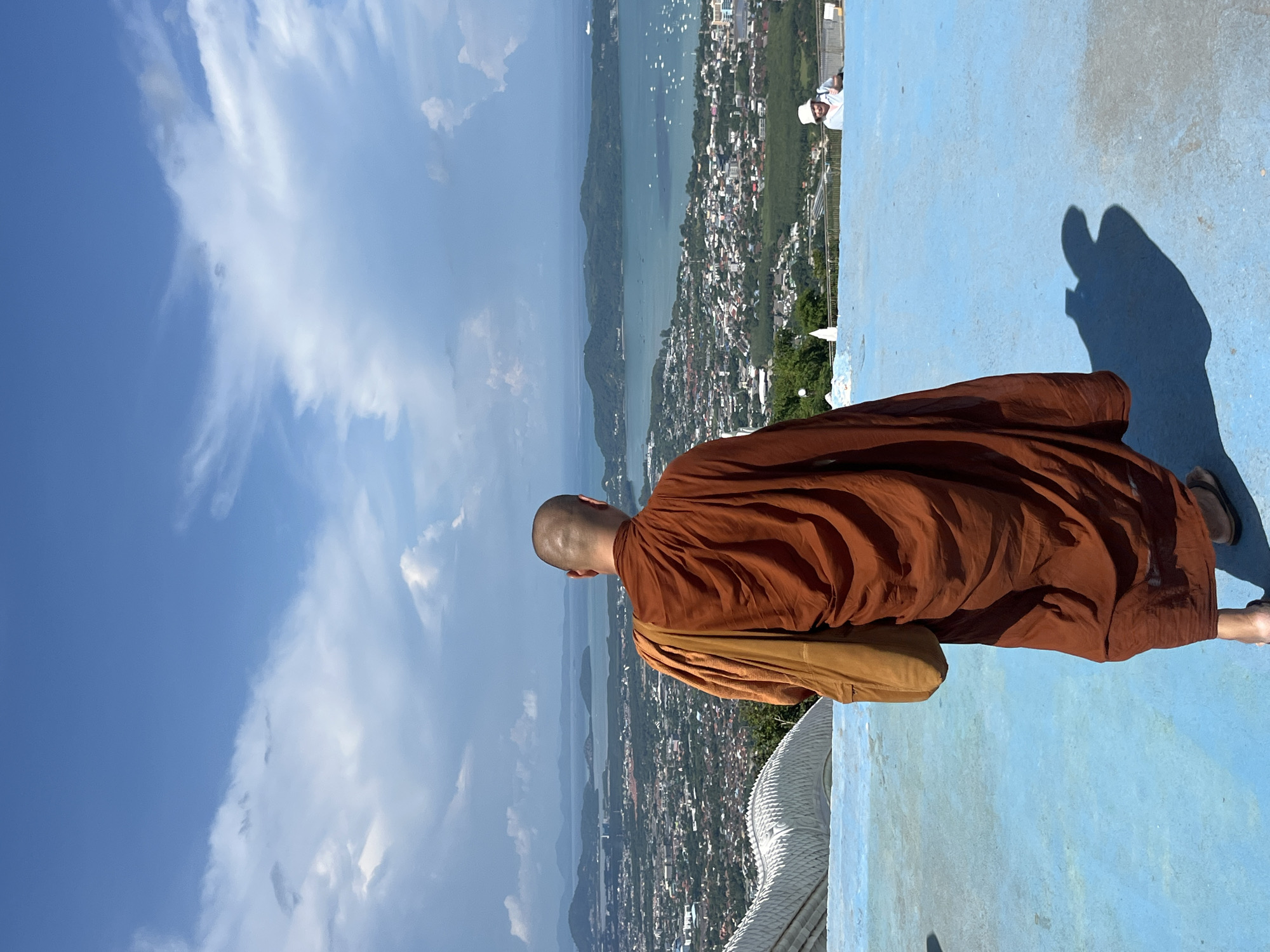 A Buddhist monk in a saffron robe stands facing away from the camera, looking out at a panoramic view of the city of Karon, Thailand. The monk has a shaved head and carries a tan bag over his shoulder. He is standing on a bright blue surface, which slopes downwards towards the city. In the distance, the city extends to meet a body of water dotted with boats, with lush green islands beyond. The sky above is a vibrant blue, with fluffy white clouds. To the right, in the mid-ground, a person wearing a white hat is visible on the blue surface. A white, serpentine structure is partially visible on the left side of the frame, suggesting the viewpoint is from an elevated temple or viewpoint. The overall scene is one of serene contemplation against a picturesque backdrop.