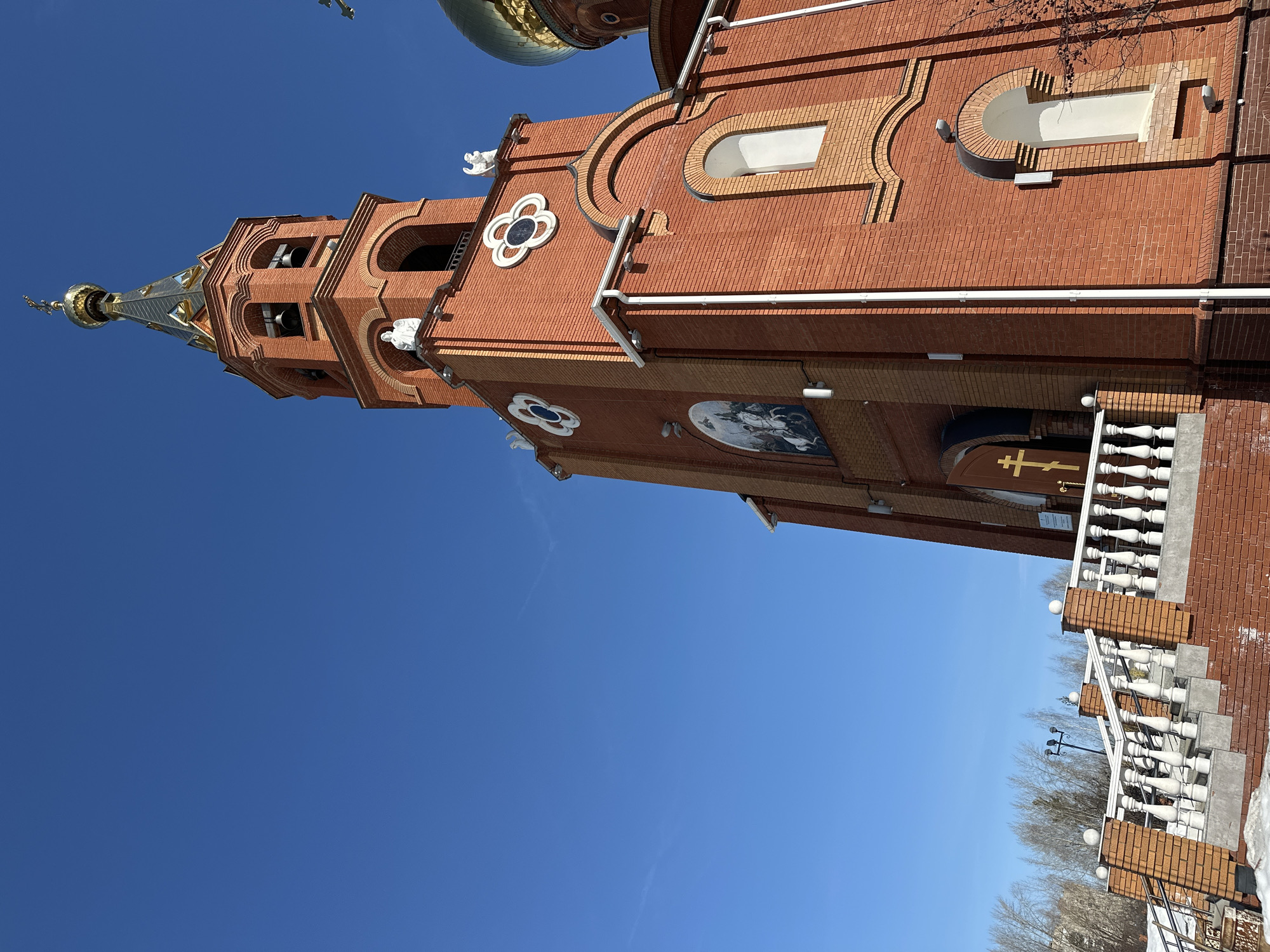 The image depicts the exterior of an Orthodox church constructed primarily from red brick, located in Chaikovsky, Russia, under a clear, deep blue sky. The prominent bell tower, also red brick, features multiple arched openings containing bells and is topped with a dark, segmented spire culminating in a golden onion dome and an Orthodox cross. White angel sculptures are positioned on ledges below the spire. The church facade incorporates lighter beige brick horizontal banding and decorative white quatrefold patterns. A large wall mural, an iconographic depiction of two figures on horseback, is visible on a section of the building. Another golden onion dome with an Orthodox cross is partially visible on the right side of the main structure. Access to an entrance involves a set of brick stairs with a white balustrade, leading to a brown door adorned with a golden Orthodox cross. Patches of snow are present on the ground adjacent to the stairs. Bare deciduous trees are visible in the background, and a streetlight stands behind the stair railing. No individuals or active interactions are observed within the scene.