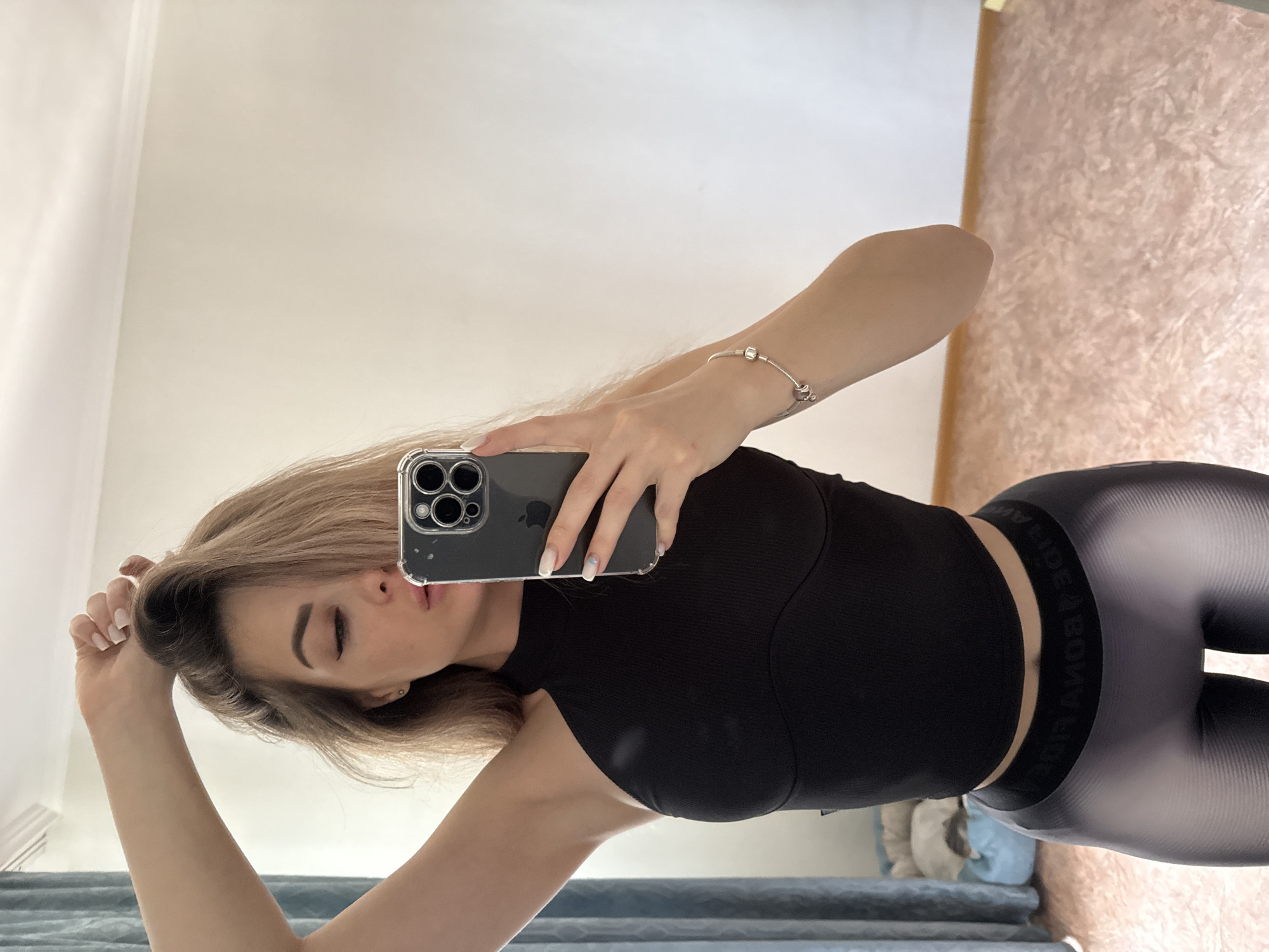 A mirror selfie shows a woman with blonde hair, wearing a black top and grey leggings with the brand name printed on the waistband. She is holding an iPhone in her right hand, and her left hand is up by her head. The background includes a plain wall and a patterned wall or possibly wallpaper. The location appears to be inside a home. The city and state are Unknown, Unknown.