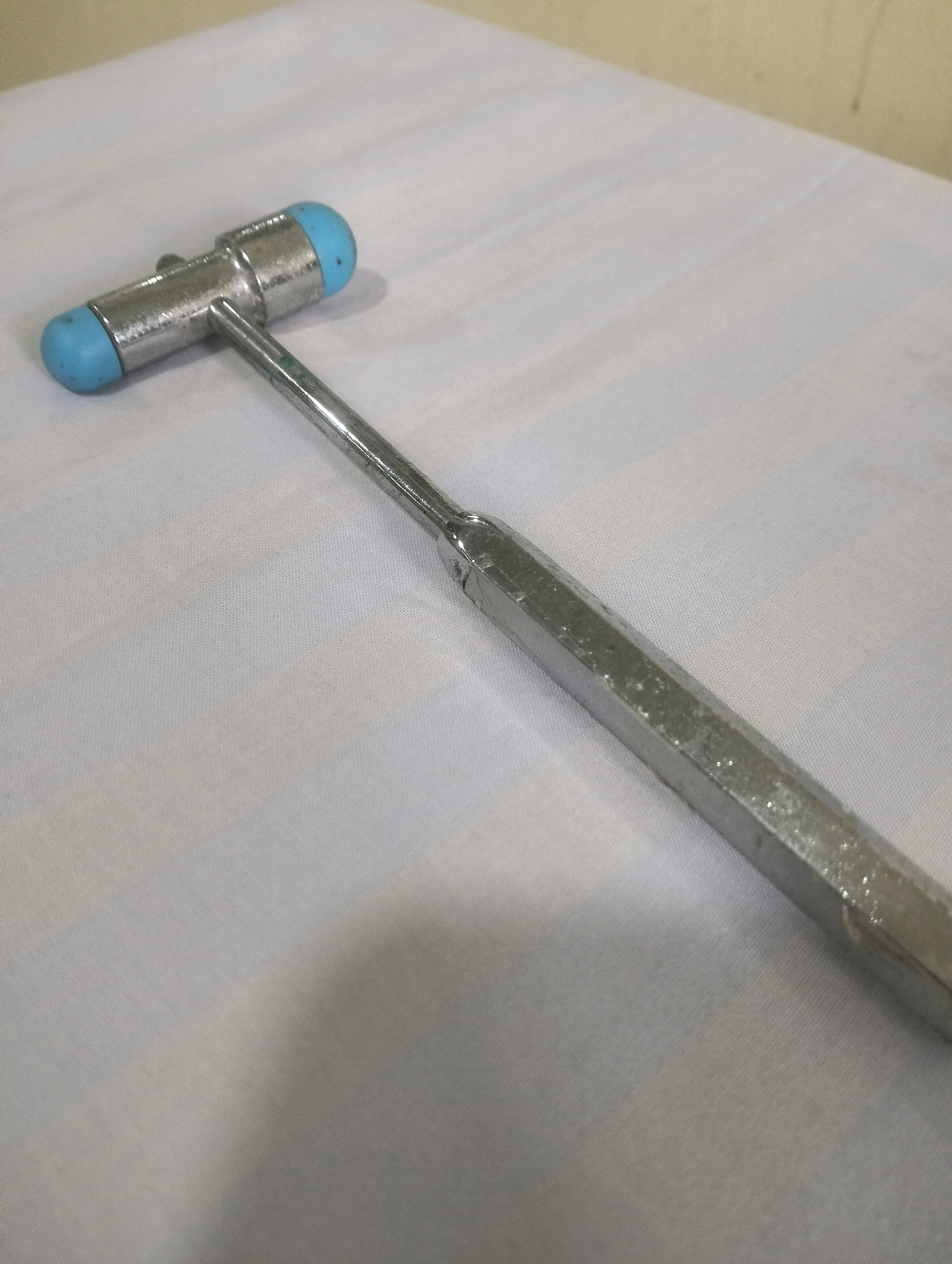 A metal reflex hammer with blue rubber ends lies on a white, textured surface, possibly a tablecloth or sheet. The hammer's handle is hexagonal and metallic, with visible marks and a textured grip. The head of the hammer consists of a metallic component with two rounded blue caps on either side, suggesting it's designed for different types of reflex testing. The background is a plain white surface with subtle folds and wrinkles, indicating it is a soft material. The lighting suggests an indoor environment, and the overall impression is of a medical or diagnostic tool placed on a surface. There is no information about the location in Air Jamban, Indonesia.