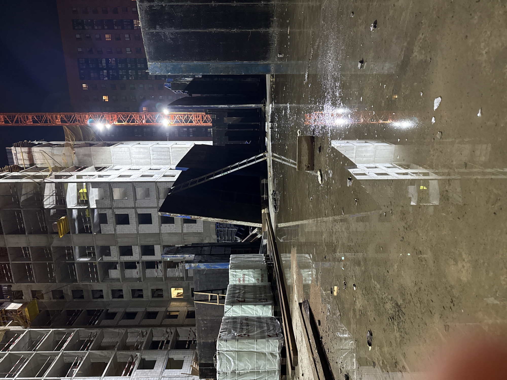 The image shows a construction site at night in Moscow, Russia. The scene is dominated by a large, unfinished apartment building, its concrete skeleton illuminated by artificial lights. A red construction crane stands tall, its boom extending towards the sky, with bright work lights attached to it. In the foreground, the ground is covered in a large puddle of muddy water, reflecting the lights and structures. Several large, white, plastic-wrapped bundles are stacked on wooden pallets to the left, likely building materials. Various construction barriers and scaffolding are visible throughout the scene. The overall impression is one of ongoing construction in an urban environment, with the water on the ground suggesting recent rain or a water source. There are no people visible in the image.