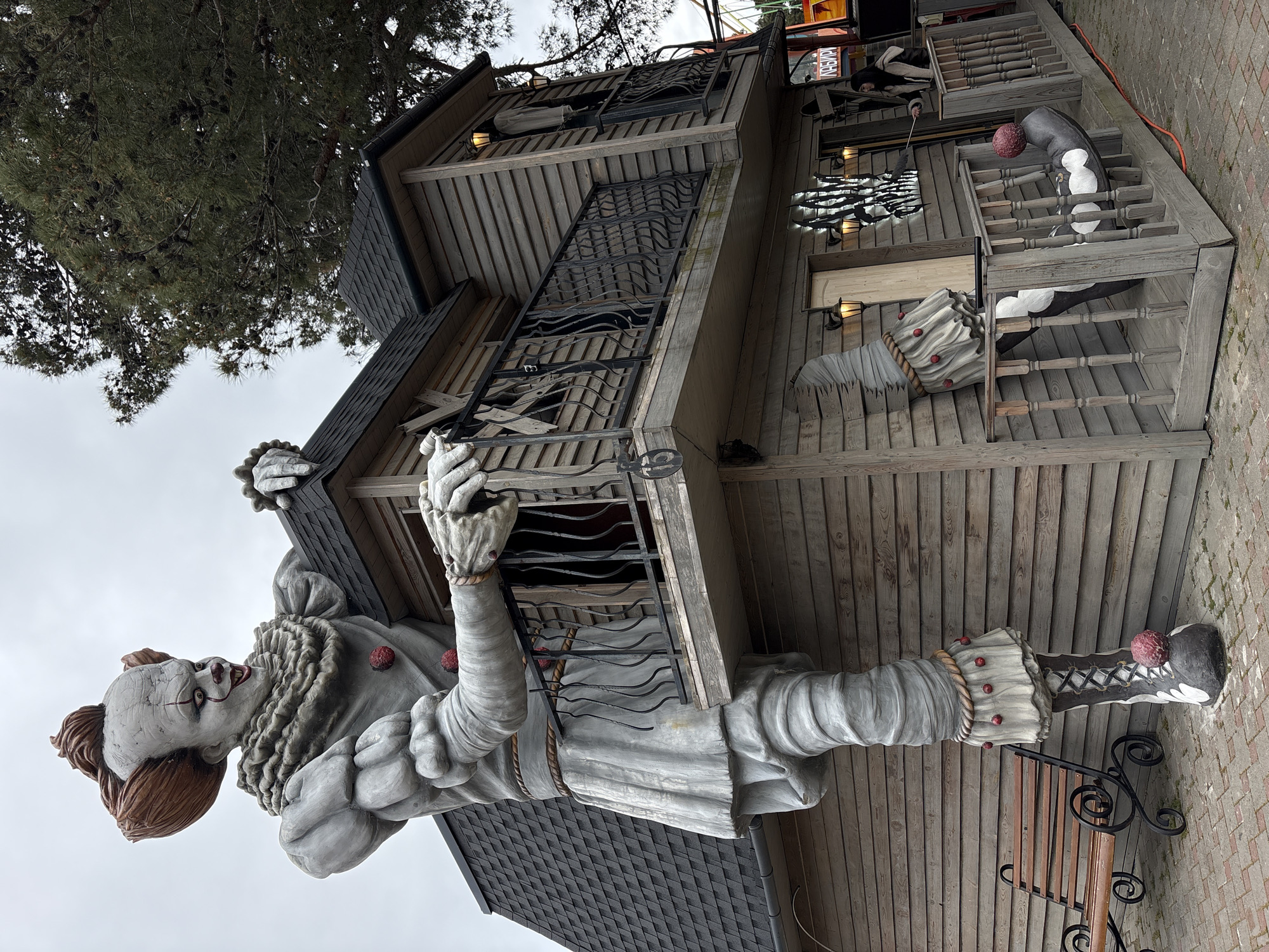 A statue of Pennywise, the clown character from Stephen King's "It", is attached to the front of a wooden three-story building with a wrought iron balcony. The building has a dark grey roof. The statue has a white face with red makeup, an orange wig, and a grey costume with red pom-poms. The location is Gelendzhik, Russia.