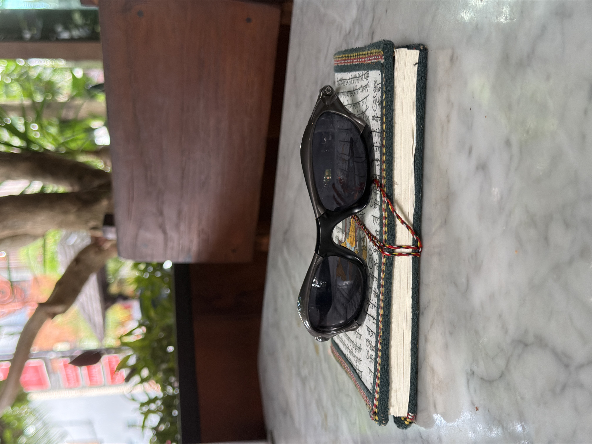On a marble table in North Kuta, Indonesia, a pair of black sunglasses rests atop a book. The book has a decorated cover with a yarn binding. In the background, a plant is visible.