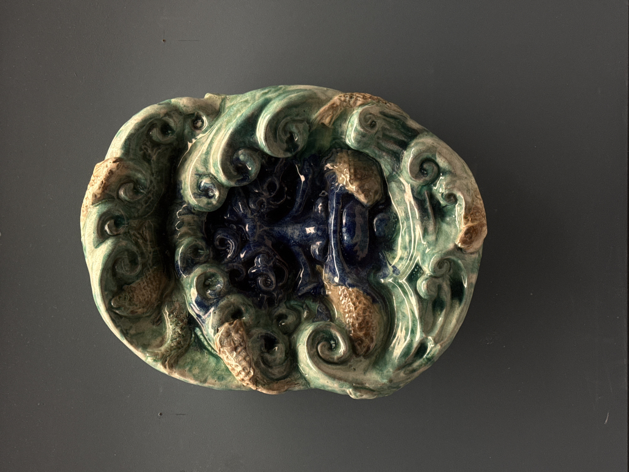A decorative ceramic plate is photographed against a plain grey background. The plate is roughly oval-shaped with a raised, sculpted design. The outer rim features stylized waves in shades of green and white, interspersed with brown, textured fish. The central area is a deeper blue with swirling patterns that create a sense of depth. The plate appears to be lying flat on the surface. The lighting is somewhat diffused, creating shadows that emphasize the texture of the ceramic. There is no indication of this image being in Moscow.