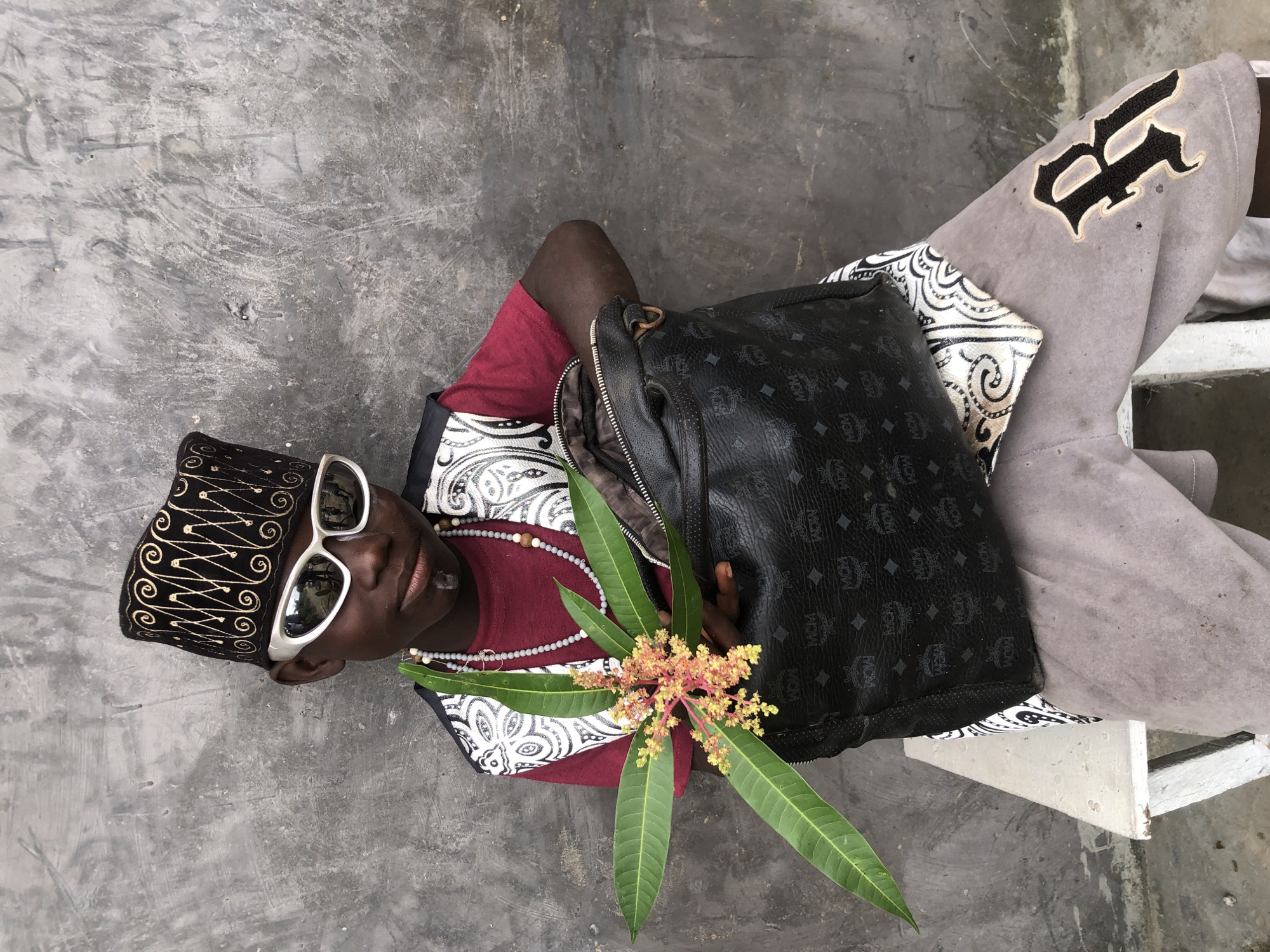 A young Black male, adorned in a black and gold patterned kufi cap and white-rimmed sunglasses, sits facing forward on a white wooden bench against a textured gray concrete wall. He wears a red t-shirt and a vest with a black and white patterned trim, along with grey pants featuring an embroidered 'R' on the right thigh. Around his neck, he wears a beaded necklace. In his left hand, he holds a green leafy branch with small pink and yellow flowers. His right hand rests on a black MCM-branded handbag. The scene appears to be outdoors, possibly in Ado, Nigeria, given the clothing style, skin tone, and the rustic background, suggesting an urban or semi-urban environment.