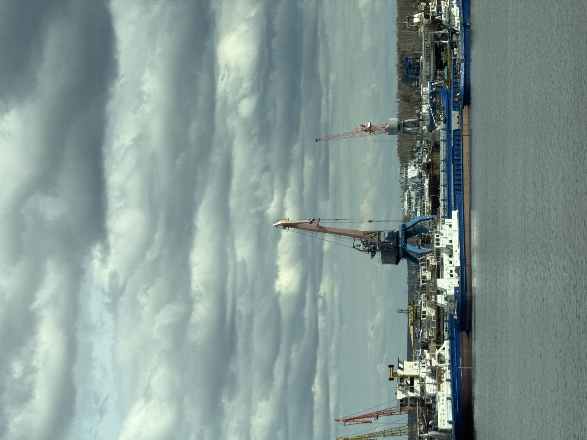 This image captures an industrial waterside scene, likely a port or shipyard, in Chaikovsky, Russia, under an overcast sky. The photograph, when correctly oriented, features a wide, calm body of water in the lower part of the frame.
Along the opposite bank, several large vessels are docked. A prominent vessel, painted blue and white, features multiple decks and industrial structures on board. Extending from the dock are several tall port cranes, suggesting ongoing cargo handling, shipbuilding, or repair activities. One crane stands out with its orange-red boom, while another features a blue framework. Further along the bank, a dense line of bare trees or dormant vegetation suggests a late autumn or winter season. Various industrial buildings and structures are visible behind the vessels and cranes.
The upper portion of the image is dominated by an expansive, cloudy sky. Large, elongated grey clouds stretch across the frame, with some lighter, brighter patches indicating diffuse daylight. No direct sunlight is visible, contributing to a muted and cool color palette throughout the scene. No people are visible anywhere in the image, and there is no discernible text on any of the vessels or structures. The overall impression is one of a quiet, working industrial environment.