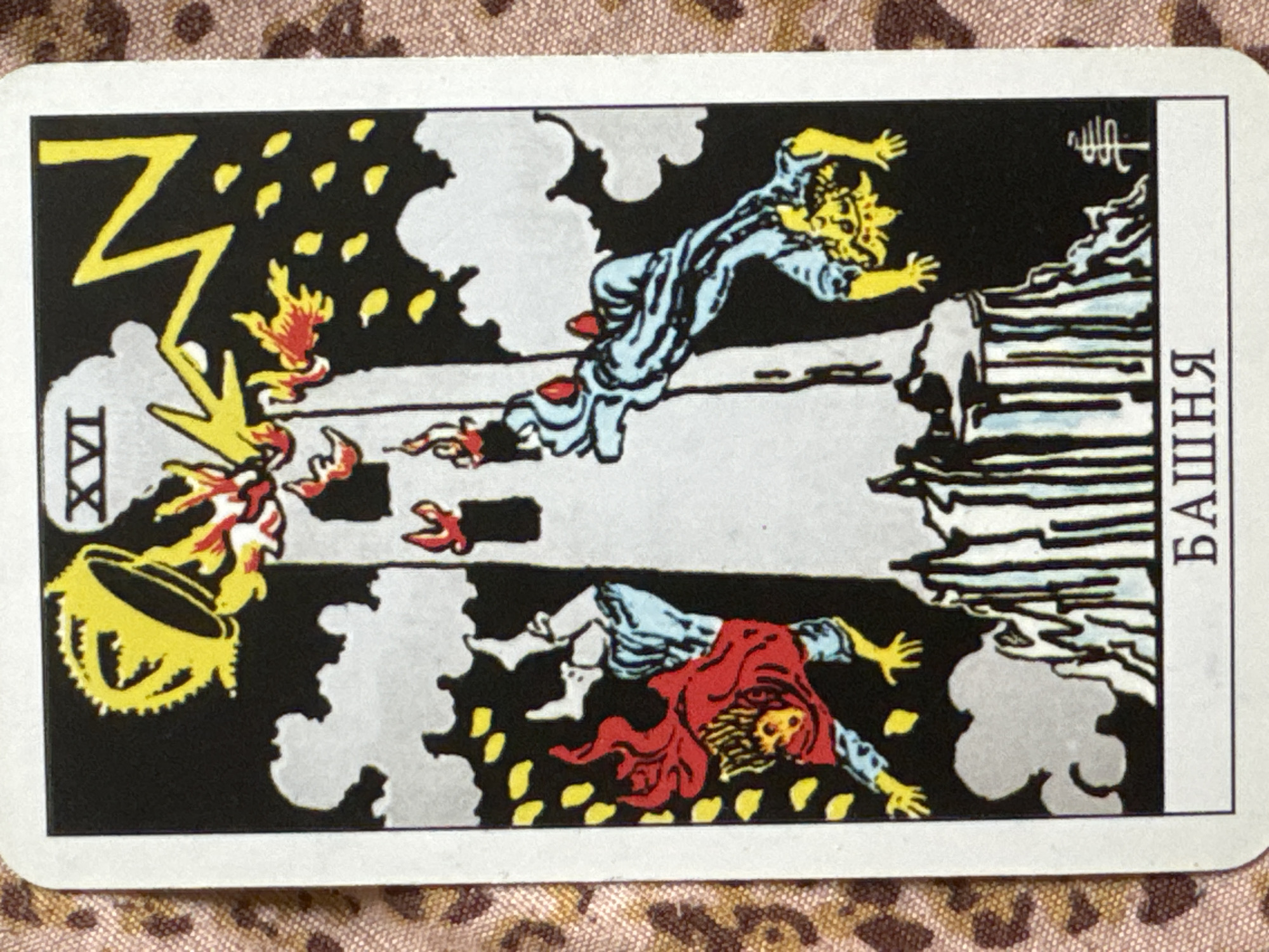 The image is a tarot card depicting "The Tower." The card features a tall, white tower being struck by a yellow lightning bolt from a stormy, black sky. The top of the tower is exploding, with flames and a golden crown falling away. Two figures are shown falling from the tower, one on the left in a red cape and blue garment, and another on the right in a blue garment with red accents. Both figures appear to be in mid-air, with expressions of distress or shock. The base of the tower is depicted as a rocky foundation, with dark, jagged shapes. Small, red flames are also visible at various points on the tower. The card has Roman numerals "XVI" at the top and a Cyrillic inscription "БАШНЯ" (Bashnya, meaning "Tower") at the bottom. There is also a small, stylized symbol resembling a tree on the right side of the card. The card is placed on a fabric with a leopard-print pattern. There is no indication that this image is located in Izhevsk, Russia.