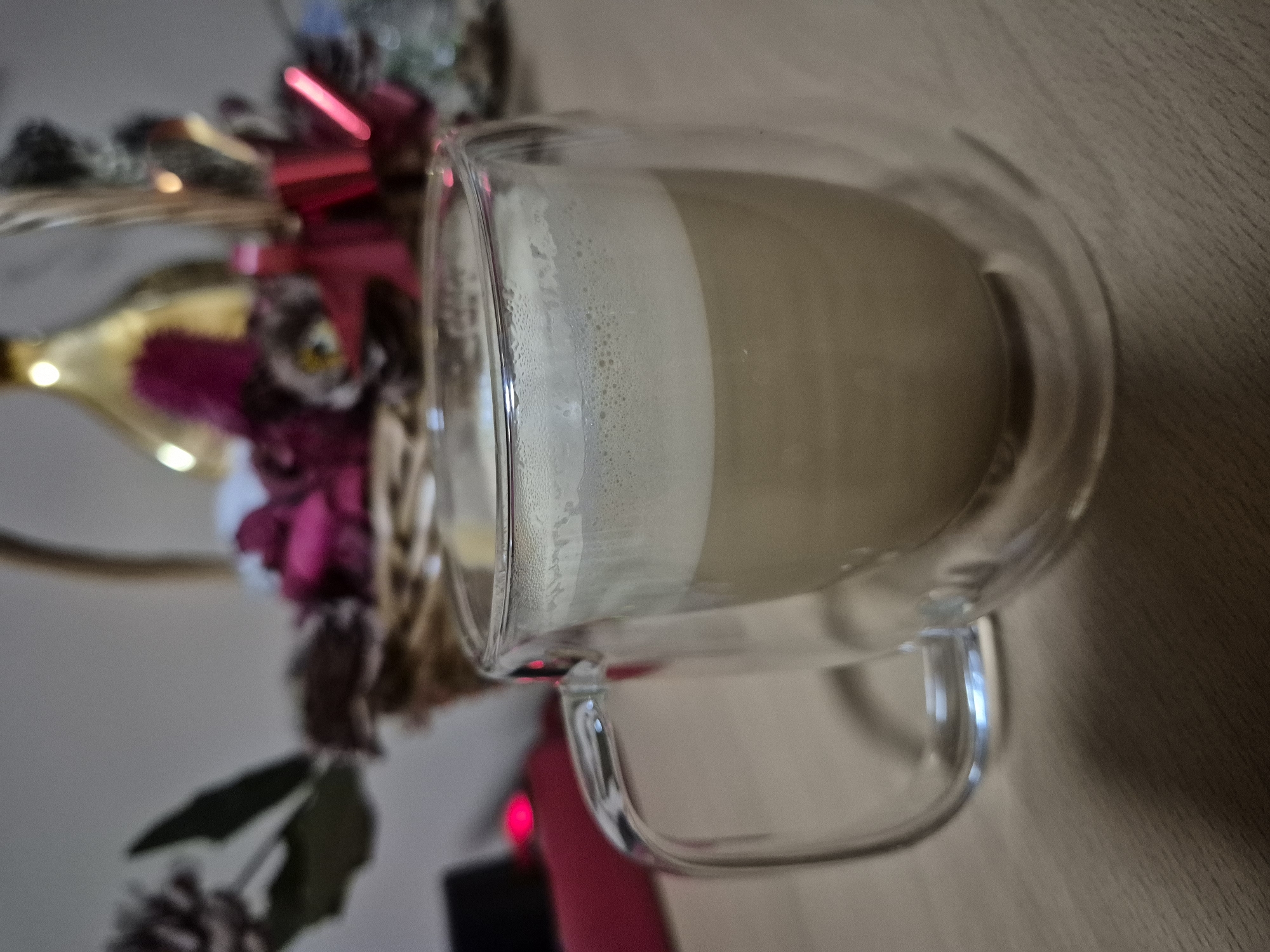 A close-up, eye-level shot captures a clear, double-walled glass mug filled with a frothy, light brown beverage, likely coffee or latte, resting on a wooden surface. The mug is positioned to the right of the frame, with its handle clearly visible. Behind the mug, a blurred background features a decorative basket containing an arrangement of red and pink flowers, pinecones, and a gold-colored bottle or vase. The lighting is dim, with some highlights reflecting off the glass and the decorative elements. The environment appears to be indoors, possibly a home or cafe setting, with no specific city or location identifiable.