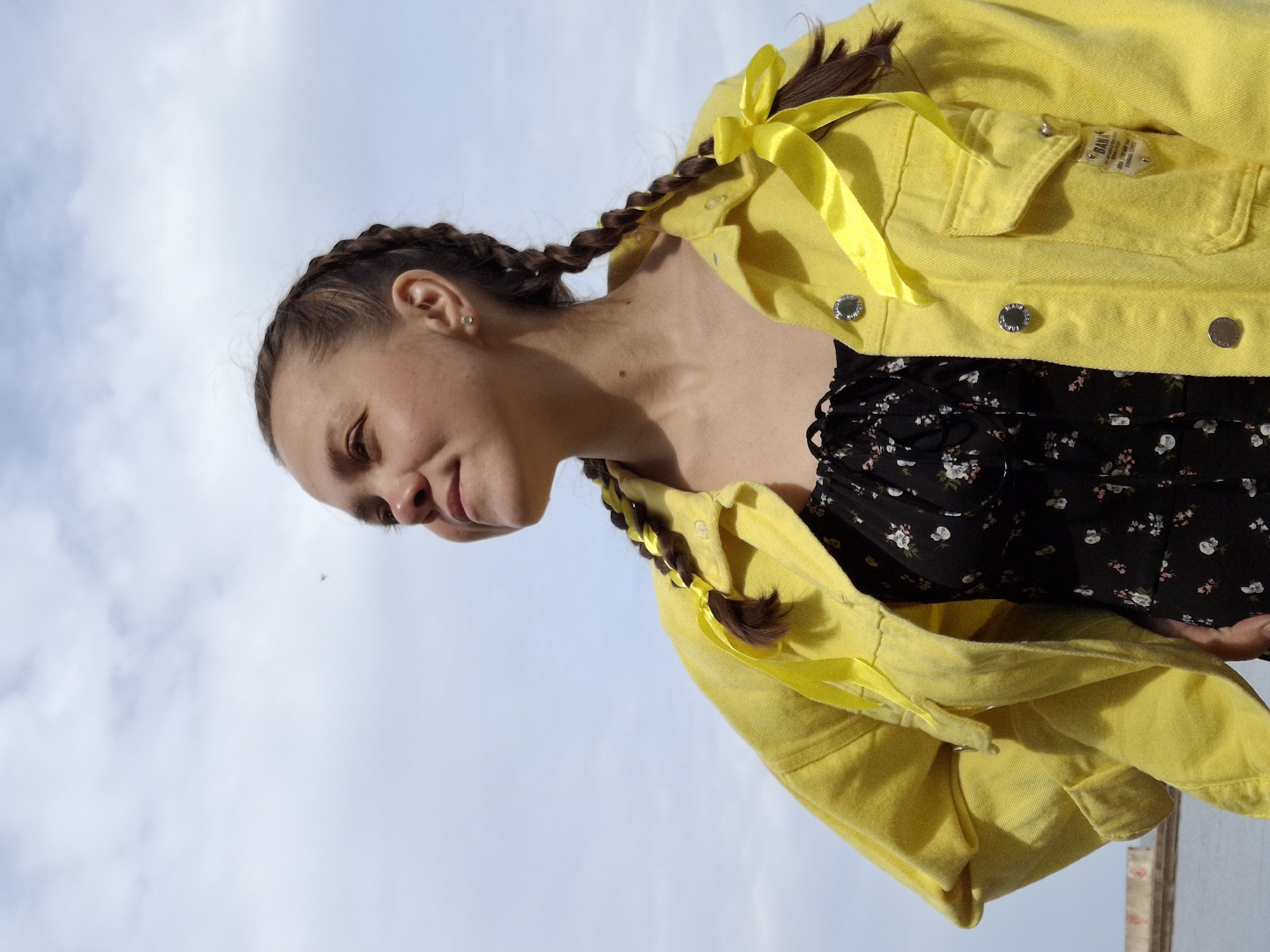A woman is seen from a low angle against a bright, overcast sky. She has dark hair braided into two plaits, each adorned with yellow ribbons. She is wearing a yellow denim jacket and a black floral print dress. Her face is turned to the side, and she appears to be looking down. The background is filled with the sky. The location is Chaikovsky, Russia.