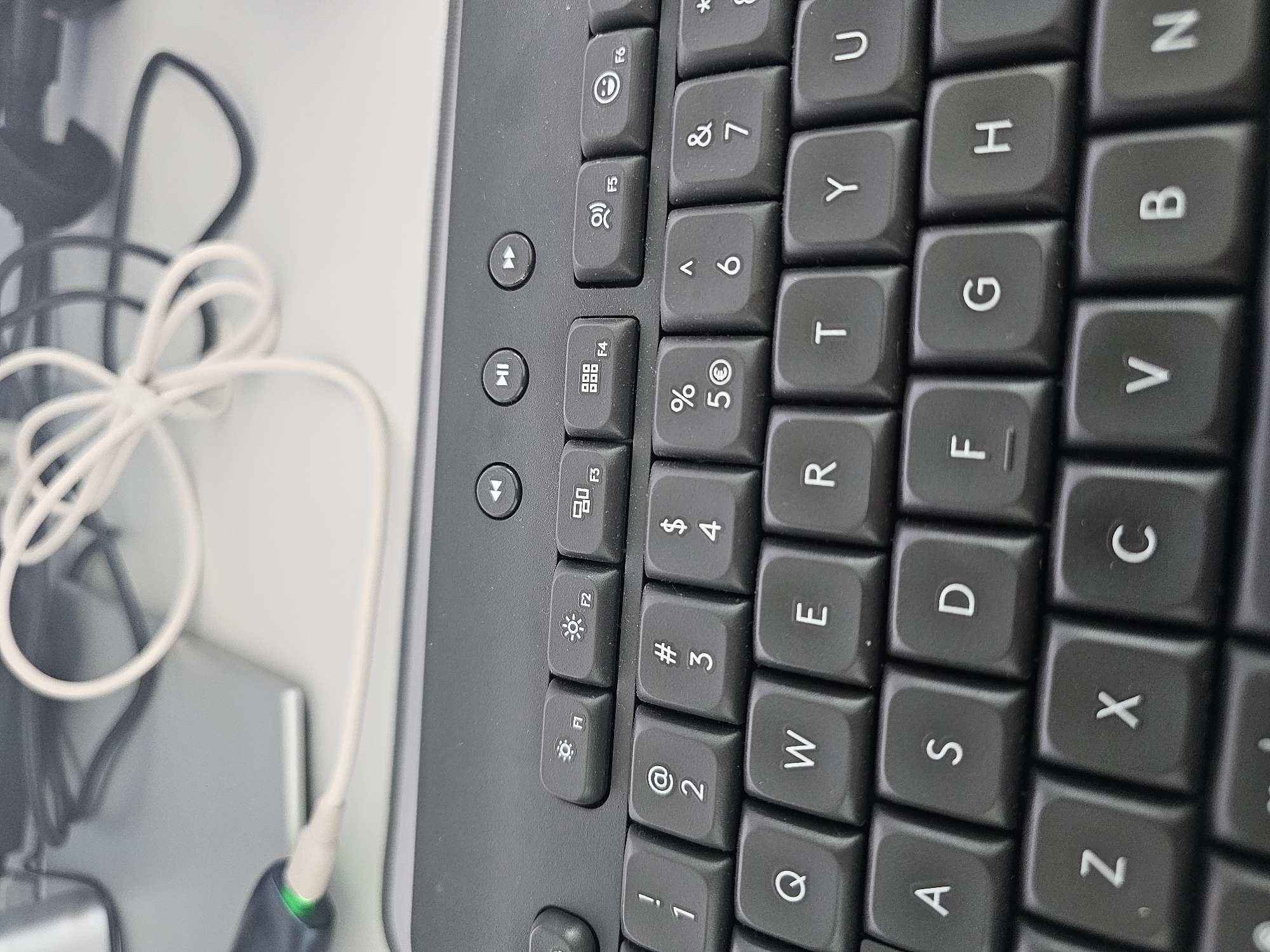 The image shows a close-up of a black keyboard and a portion of a laptop. A white cable is plugged into a green and black device, which is then connected to the laptop. The cable is loosely coiled on the desk. The keyboard has standard QWERTY layout. Function keys are visible at the top, labeled F1 through F6. The environment appears to be an office or home workspace. There are no people visible in the image. The location in the city of Bozhurishte, Bulgaria, is not discernible from the image itself.