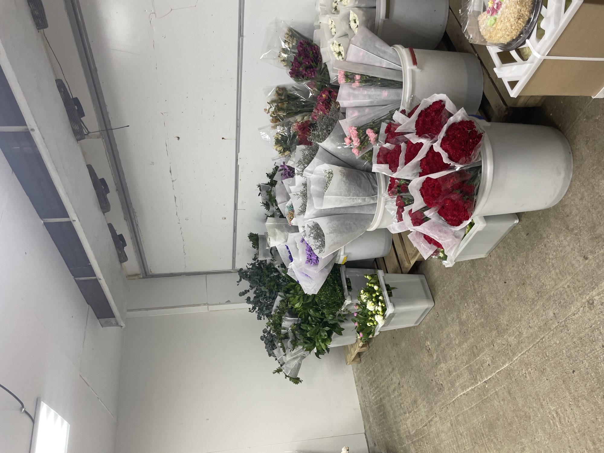 The image depicts an indoor setting, likely a floral warehouse or cold storage area, containing numerous buckets and containers filled with cut flowers and greenery. The flowers are varied in color and type, including reds, pinks, purples, whites, and greens. Many of the flower bouquets are wrapped in clear plastic and white paper. The containers are made of white plastic and appear to be sitting on wooden pallets. The environment consists of a white wall with air vents, a white ceiling with light fixtures, and a concrete floor. The image does not contain any information about the city of Izhevsk, Russia.