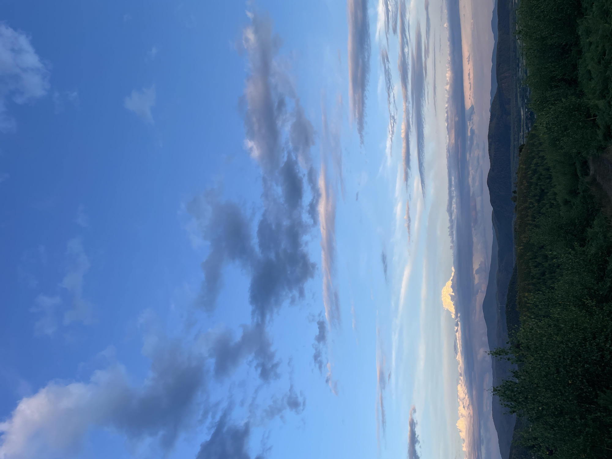 The image depicts a landscape view from a high vantage point overlooking rolling hills and distant mountains. The sky is a vibrant blue, filled with scattered clouds of varying sizes and textures. Some clouds are dark and ominous, while others are wispy and illuminated by the setting sun, casting a soft pink and orange hue. In the foreground, lush green trees and foliage create a dense canopy, obscuring the ground. The midground reveals a series of undulating hills covered in a mix of trees and vegetation. Further in the distance, layered mountain ranges extend towards the horizon, their peaks softened by the atmospheric haze. A prominent, brightly lit cloud formation stands out against the darker clouds on the horizon, resembling a colossal cauliflower. The overall scene suggests a serene natural environment during the late afternoon or early evening. The location is identified as Rokosovo, Ukraine.