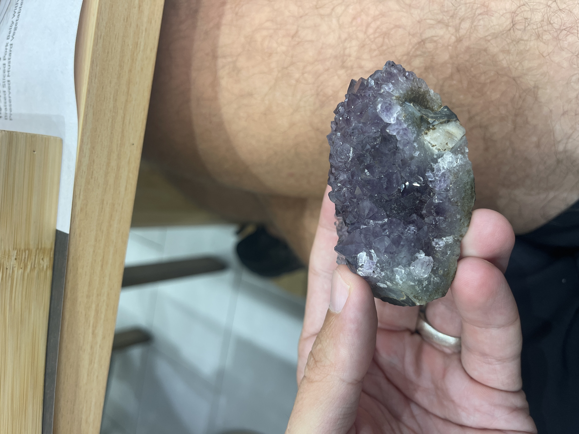 A person's hand, adorned with a silver ring on their left index finger, holds a raw amethyst geode. The geode displays a cluster of deep purple crystals, some of which are translucent and catch the light. The background is out of focus, showing the texture of a person's leg and a wooden surface, likely part of a table or desk. The scene suggests an indoor environment, but the specific location within Kuala Lumpur, Malaysia, cannot be determined from the visual information provided. The lighting is natural and appears to be coming from above.