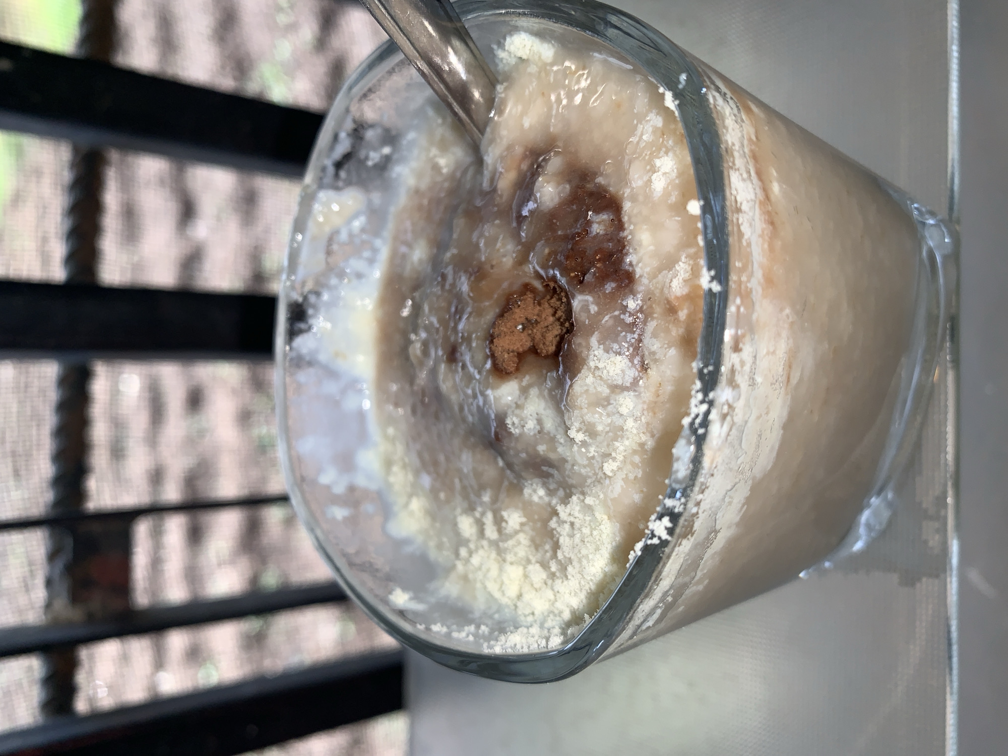 A glass containing a light brown smoothie or shake with a spoon inside is shown. The liquid appears to have a powdered substance sprinkled on top, with brown specks mixed in. The glass is positioned on a surface with a shiny appearance. Outside, there are dark horizontal bars with blurred vegetation behind them. The location in the city of Rafin Par, Nigeria cannot be determined from the image.