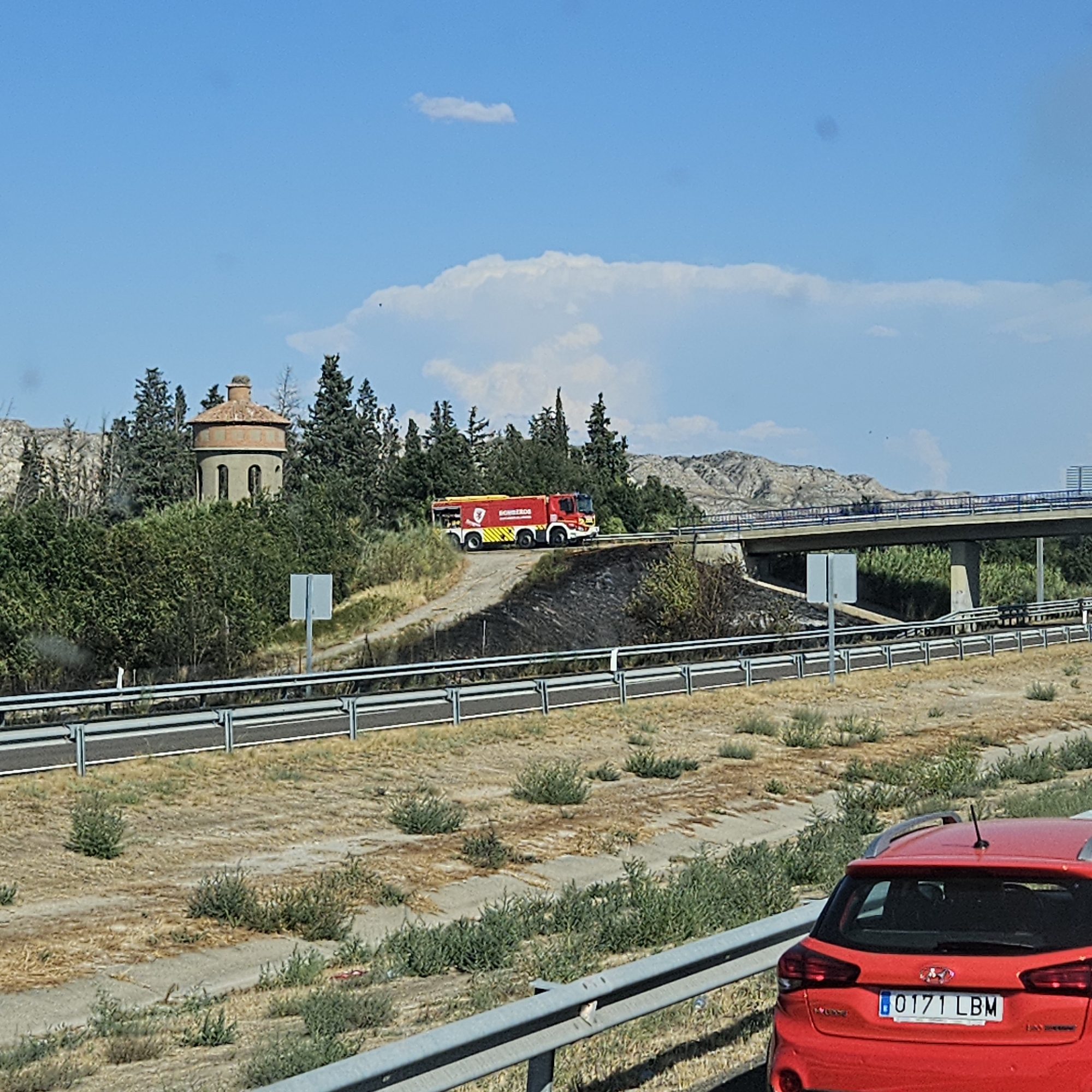 In Villafranca de Ebro, Spain, on a clear, sunny day with scattered clouds, a red and yellow fire truck is driving on a dirt road that leads to a highway overpass. The fire truck, marked with "BOMBEROS," appears to be responding to a smoldering brush fire visible on the embankment next to the road. In the background, a circular stone tower with a red tiled roof stands amidst trees, and a multi-lane highway overpass stretches across the scene. The surrounding landscape is dry and characterized by sparse vegetation, with a red car partially visible in the foreground.
