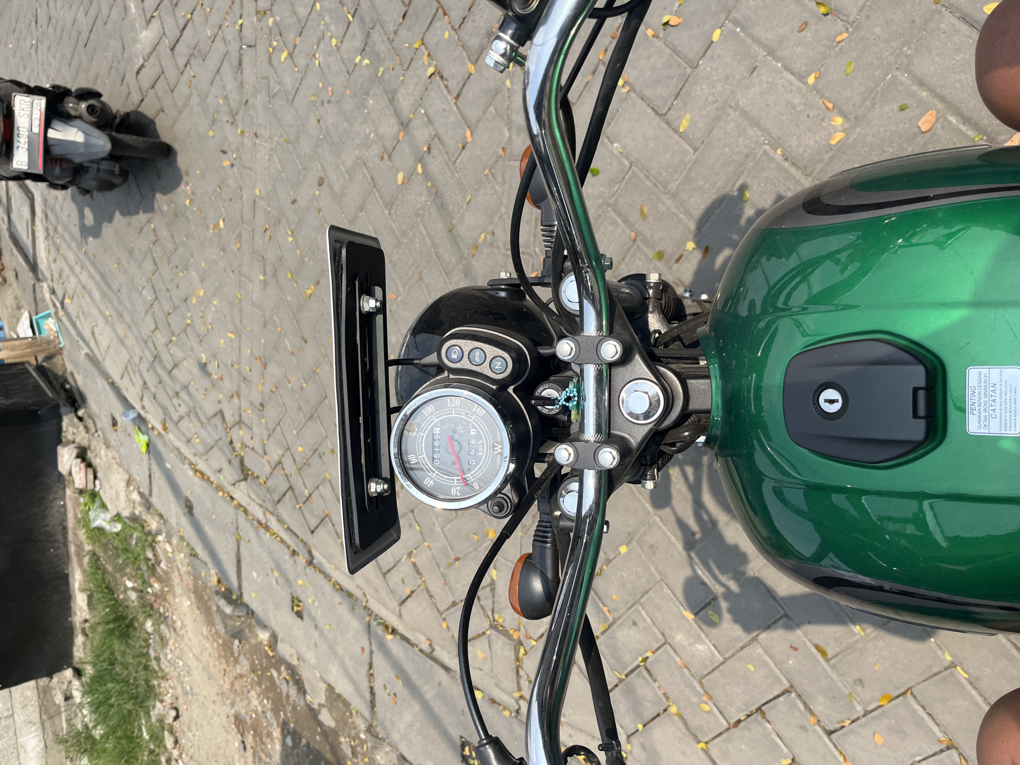The image is a first-person perspective shot from a green motorcycle parked on a paved walkway in Tangerang, Indonesia. The motorcycle's handlebars, dashboard with a speedometer, and part of the fuel tank are visible. In the background, another motorcycle is parked, and foliage is scattered on the ground. The lighting suggests it is daytime.