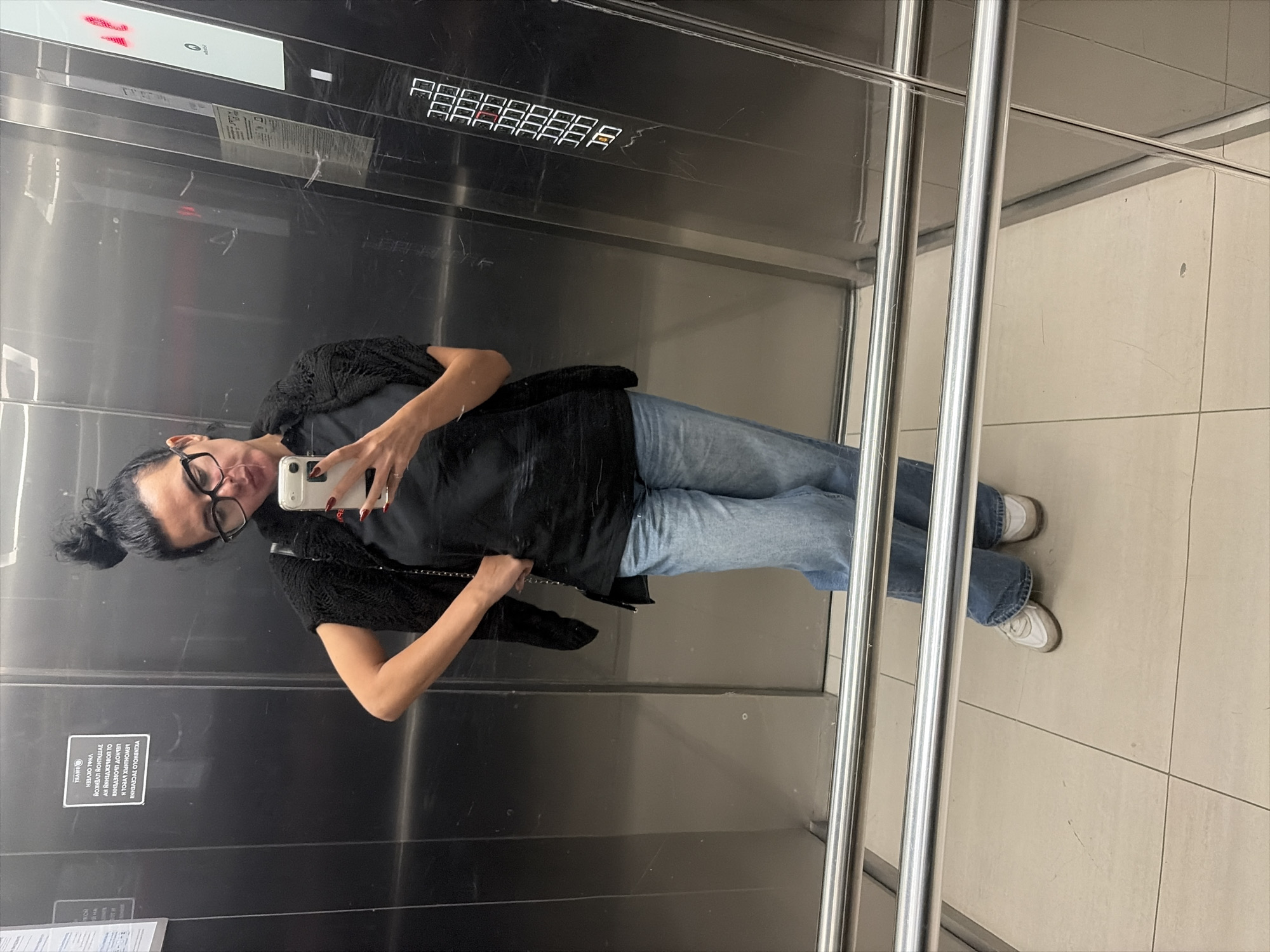 A woman with her hair in a bun and wearing glasses takes a selfie in an elevator. She is wearing a black t-shirt, a black fuzzy vest, light blue jeans, and white sneakers. The elevator has stainless steel walls and a tiled floor. There are reflective surfaces and elevator buttons visible. The scene appears to be indoors, likely in a public building.